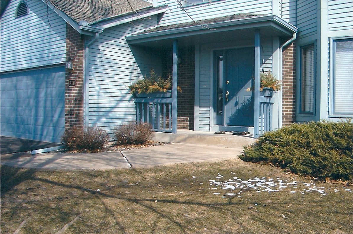 Blue house with porch, shrubs, and patchy grass.