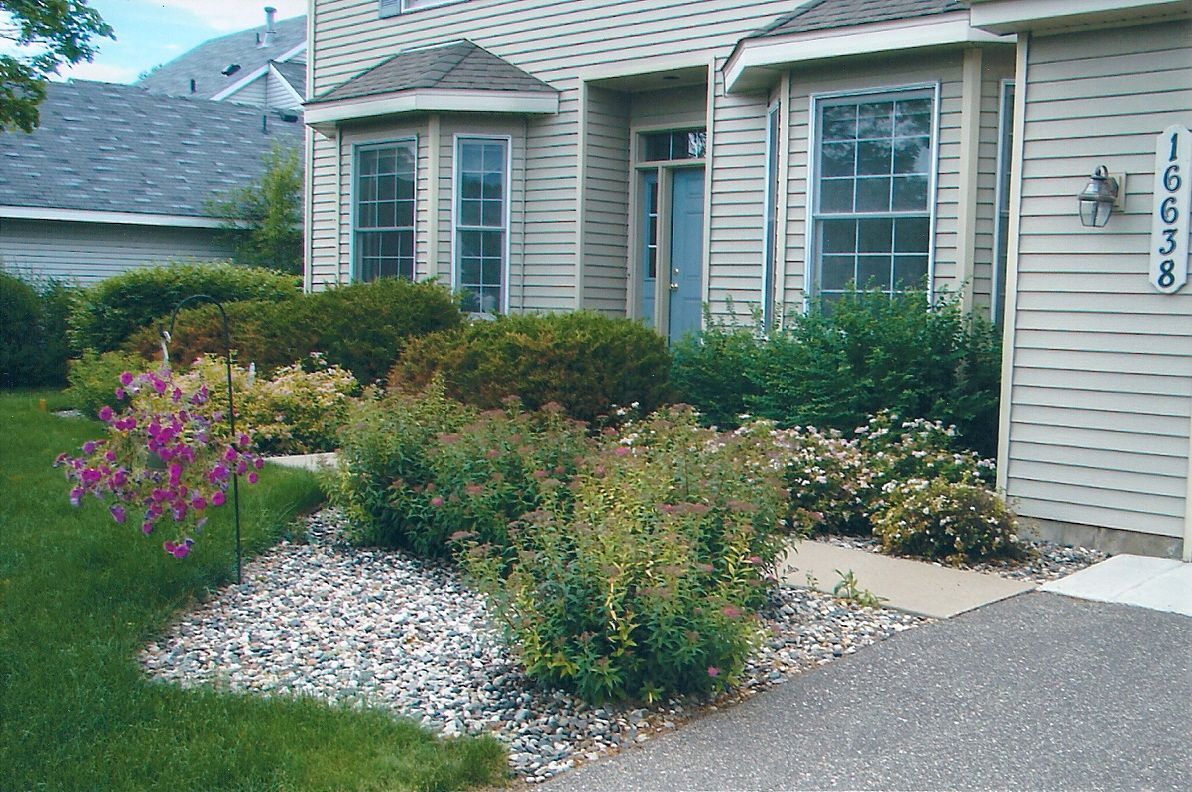 House front with a gravel and plant garden bed. Green shrubs, pink flowers, and a paved walkway.