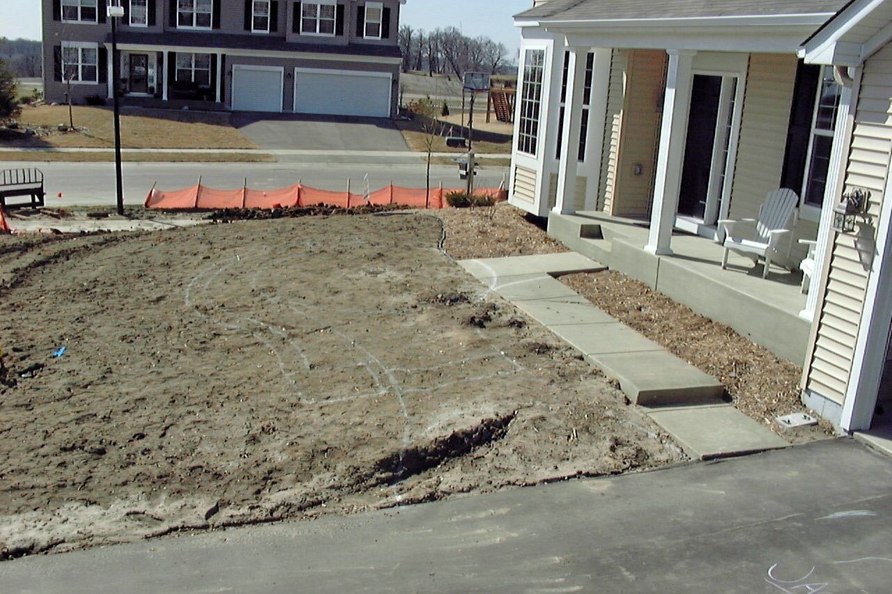 A house with a porch and driveway. Dirt yard in front, and orange safety fencing.
