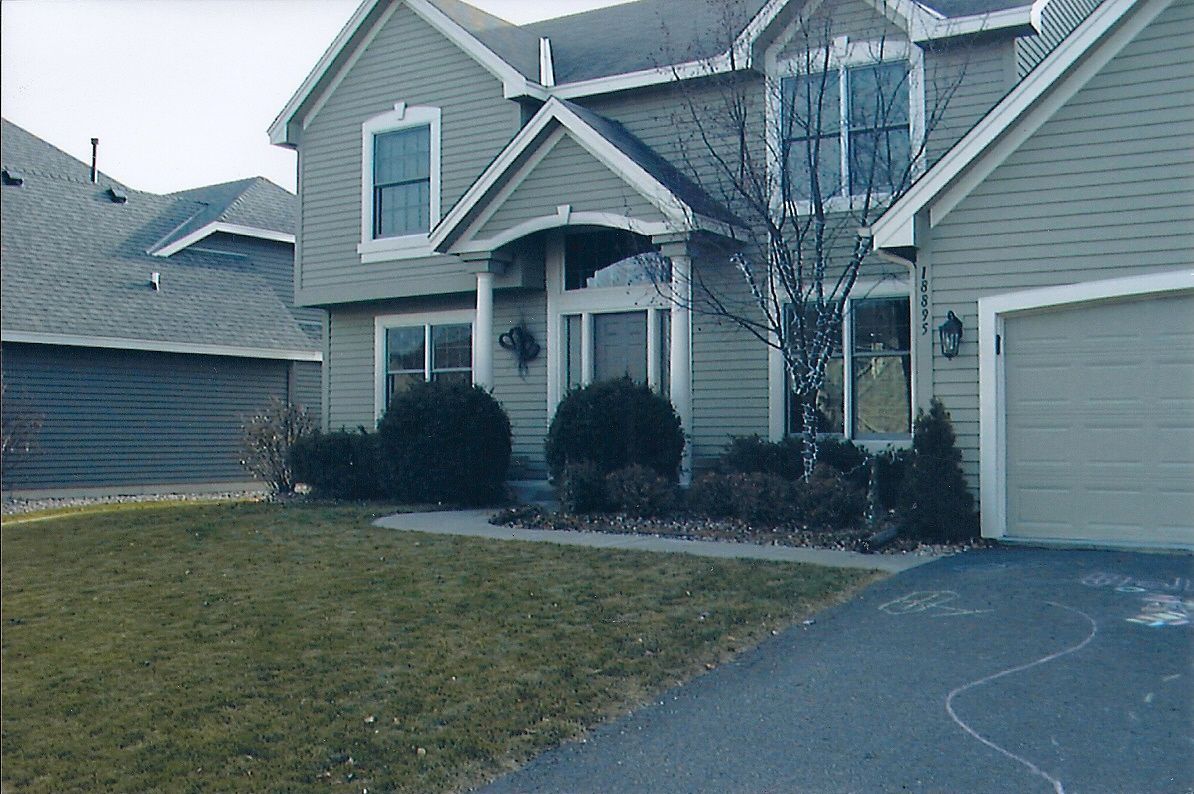 Two-story house with light green siding and a gray driveway. Front yard with bushes and a bare tree.