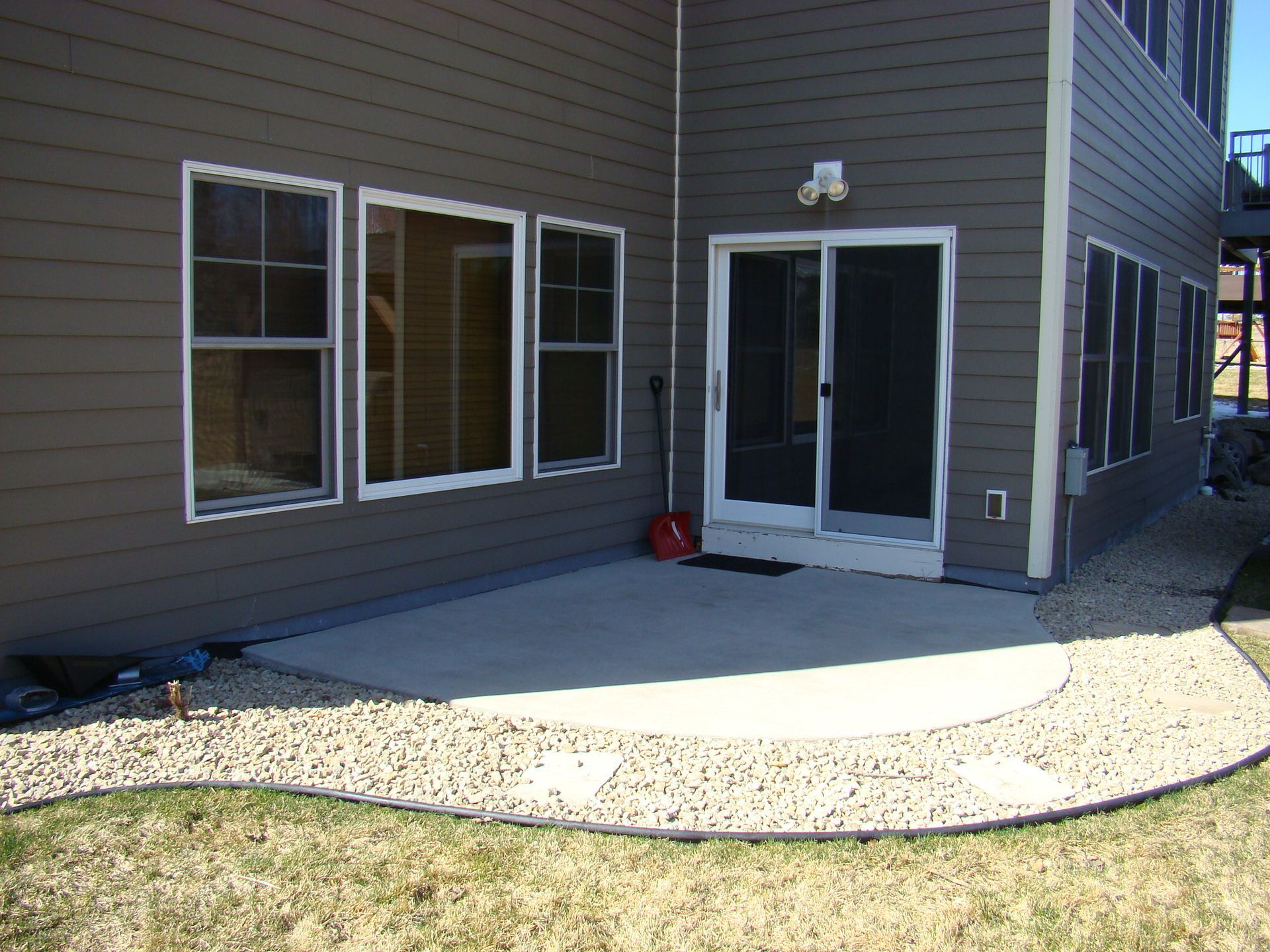 Back patio with concrete surface, brown siding, windows, and sliding door. Gravel border surrounds the patio.