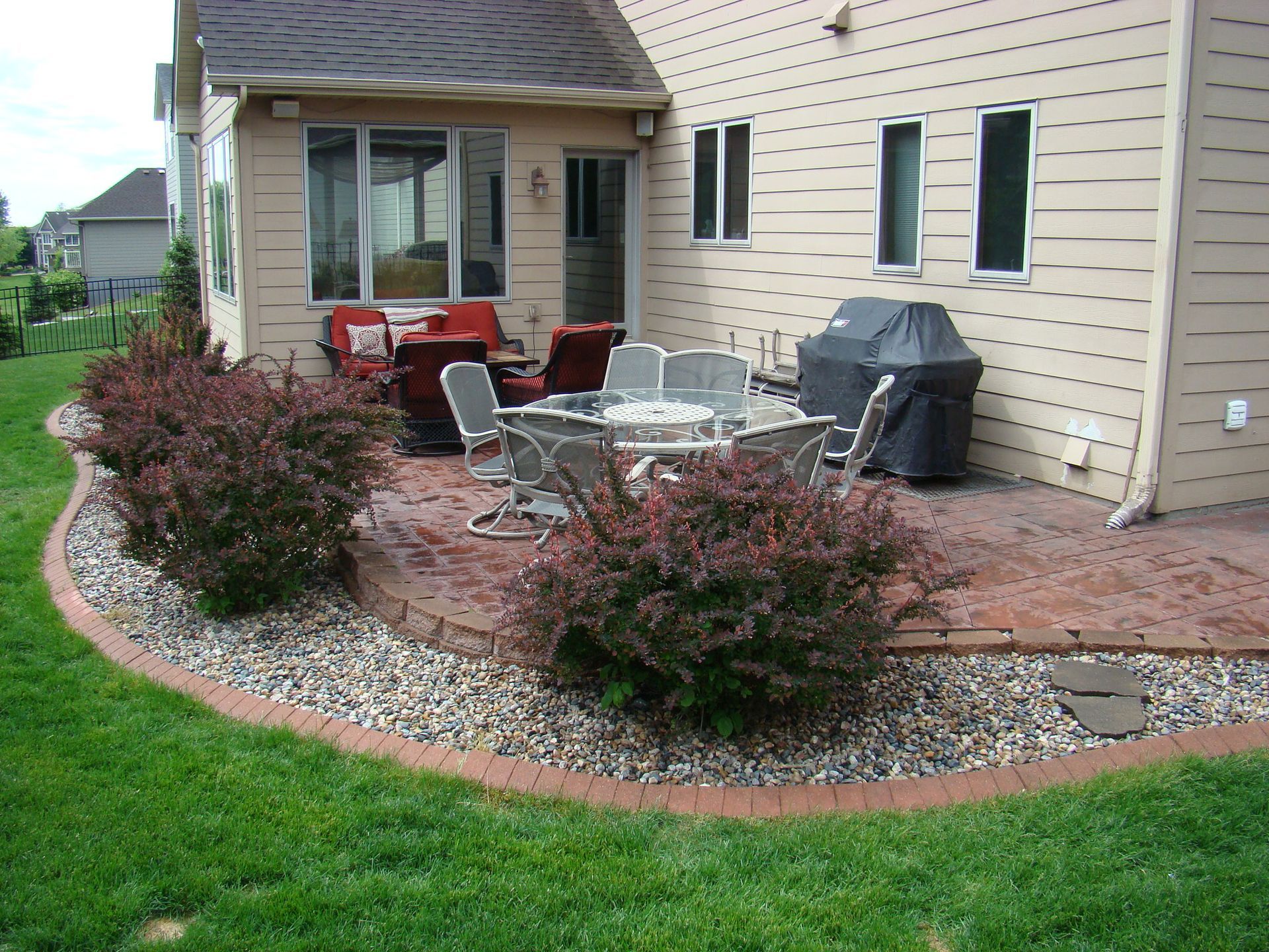 Backyard patio with red brick border, gravel, shrubs, and outdoor furniture.