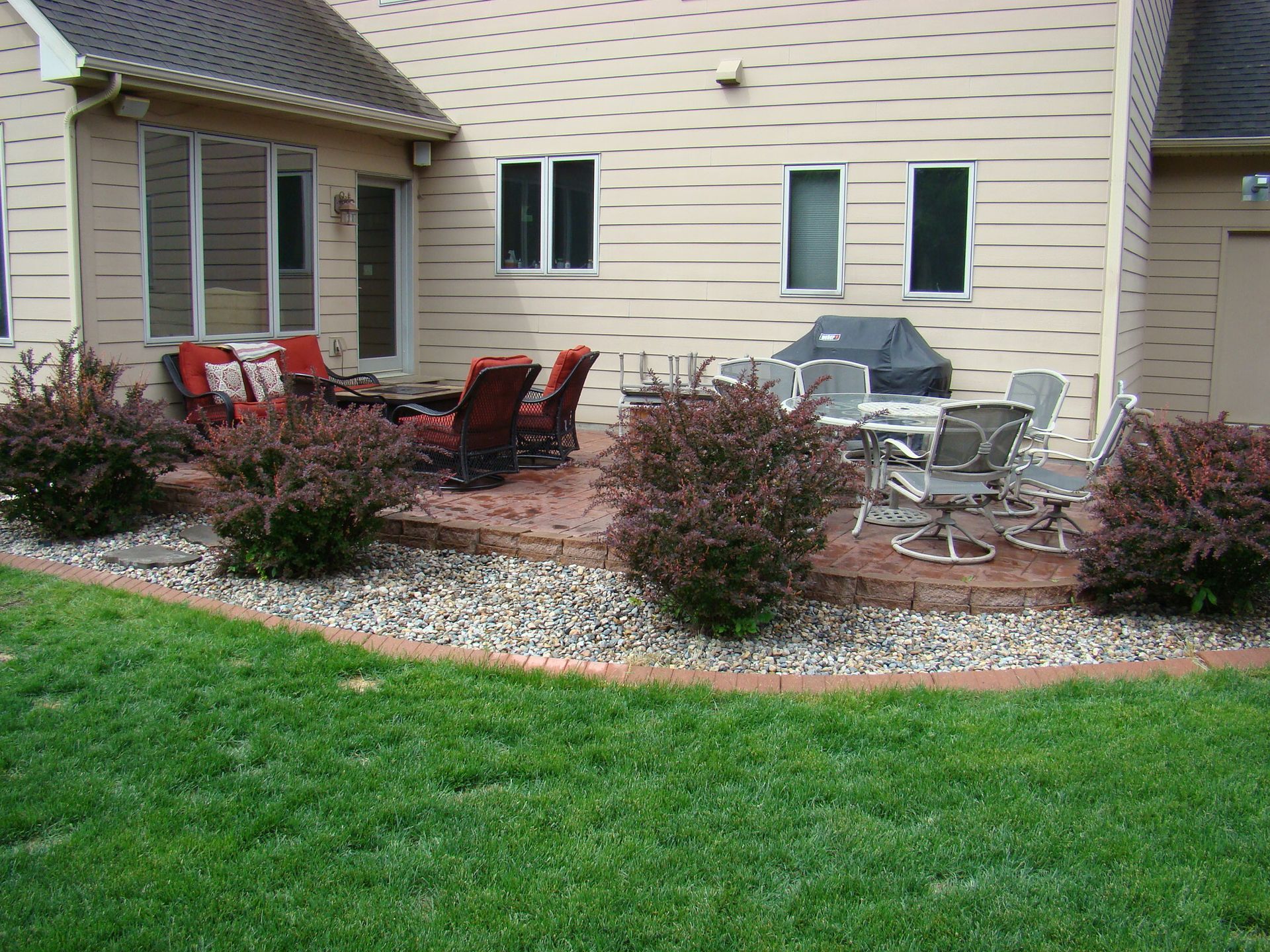 Patio with outdoor furniture, shrubs, and gravel border next to a house with windows. Green lawn.