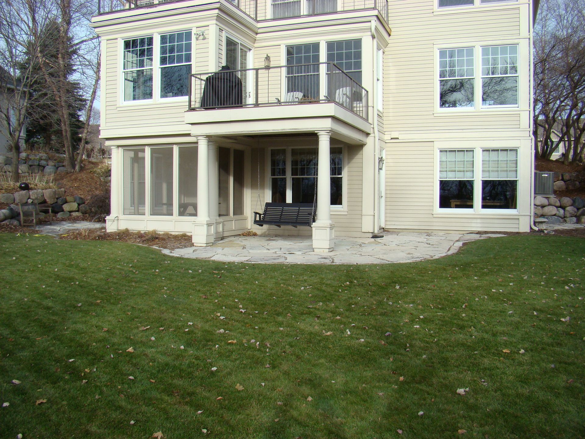 Back of a house with a screened porch and a balcony. Beige siding, green lawn, and a stone patio.