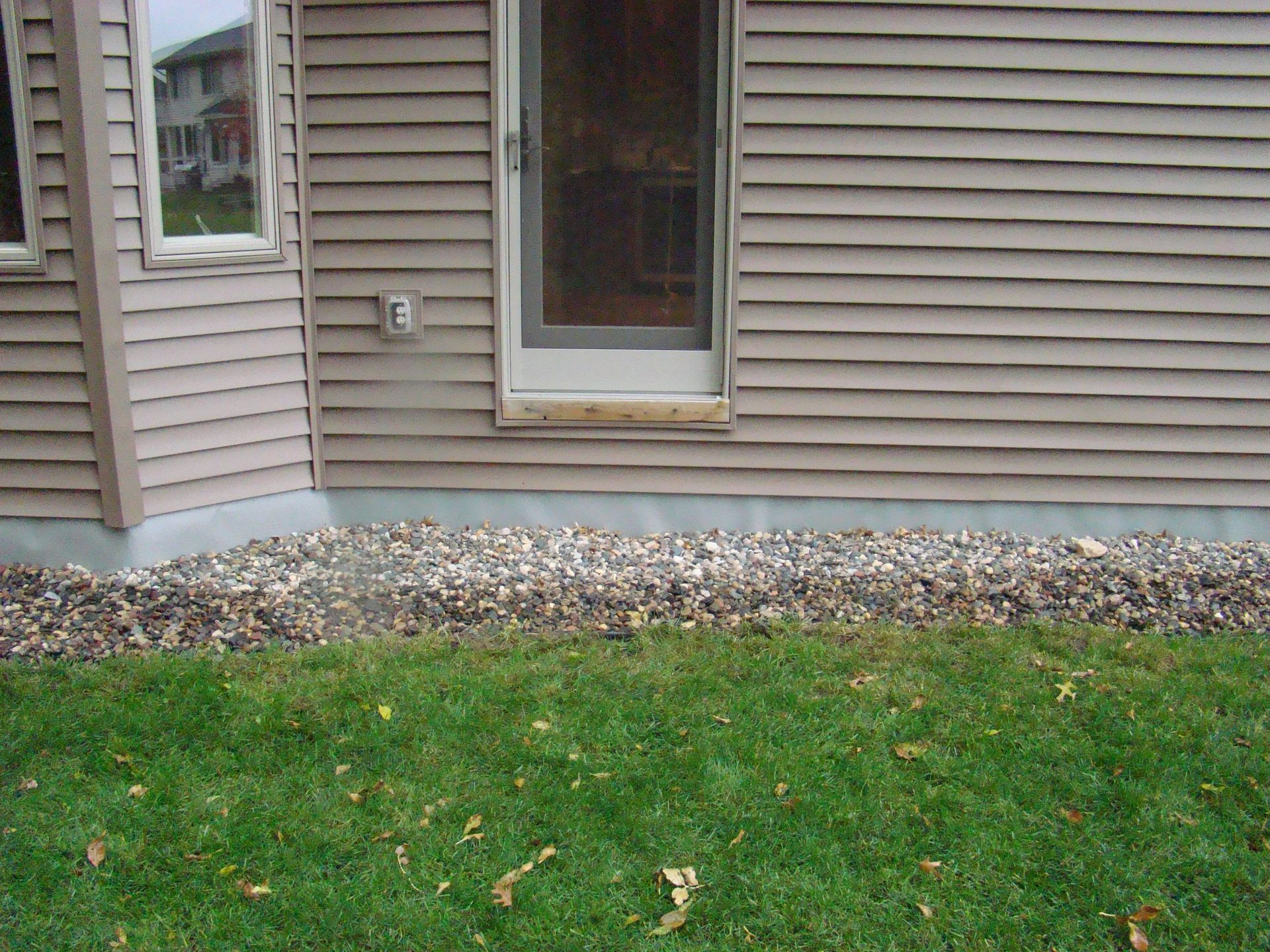 Tan siding, door, and window on a house. Gravel bed borders the foundation, green grass in the foreground.