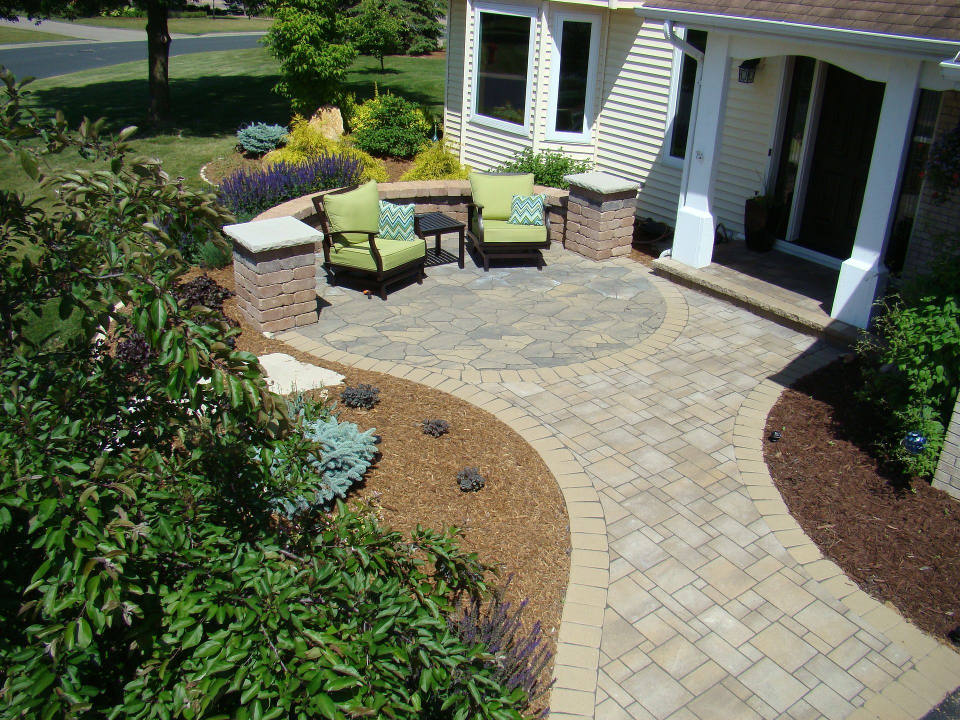 Brick patio with seating area; beige and brown brickwork, green chairs.