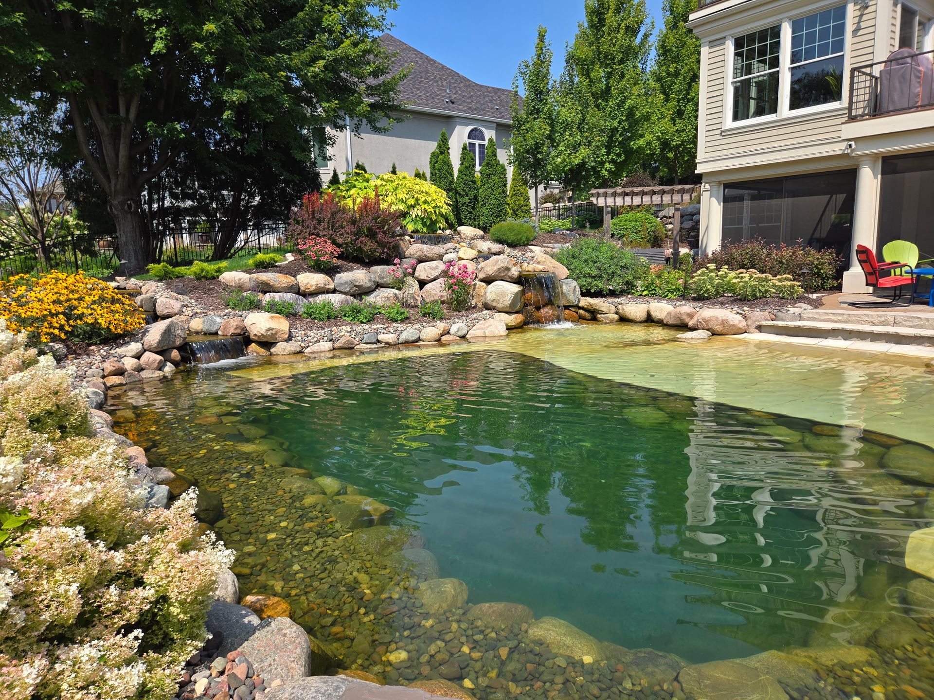 A landscaped backyard with a pond, waterfall, and colorful plants near a house under a blue sky.