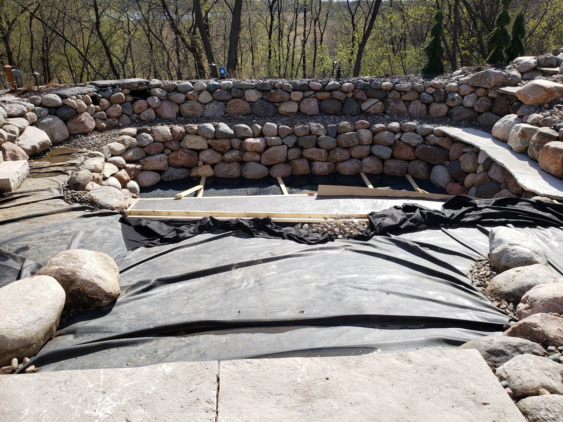 Stone-lined pond under construction, black liner visible. Wooden beams, rocks, and walkways in a natural setting.