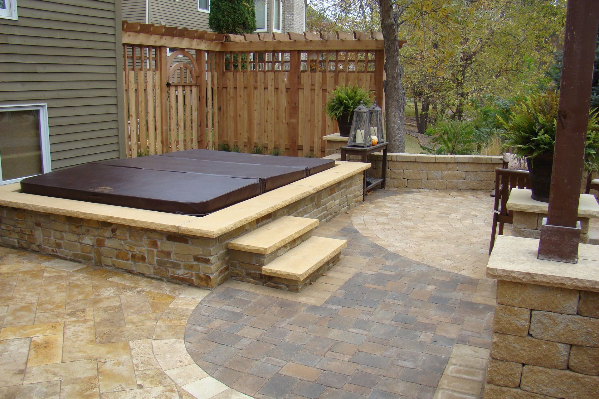 Stone patio with hot tub on a raised stone platform with steps, wooden fence, and landscaping.