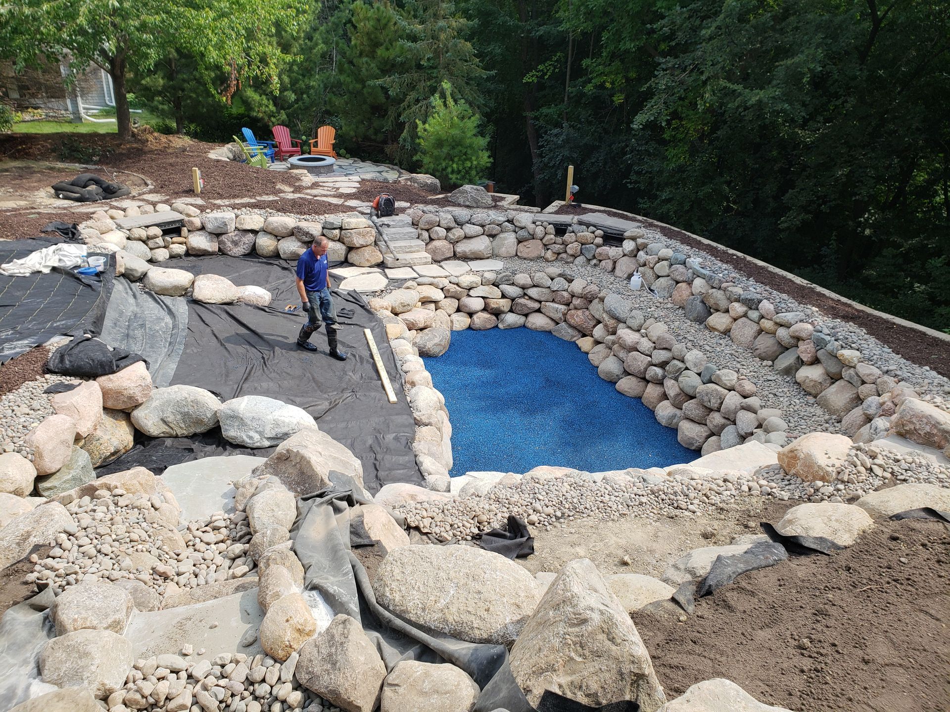 A partially built water feature with a man working. Rocks border a blue-lined pond, surrounded by more rocks and dirt.