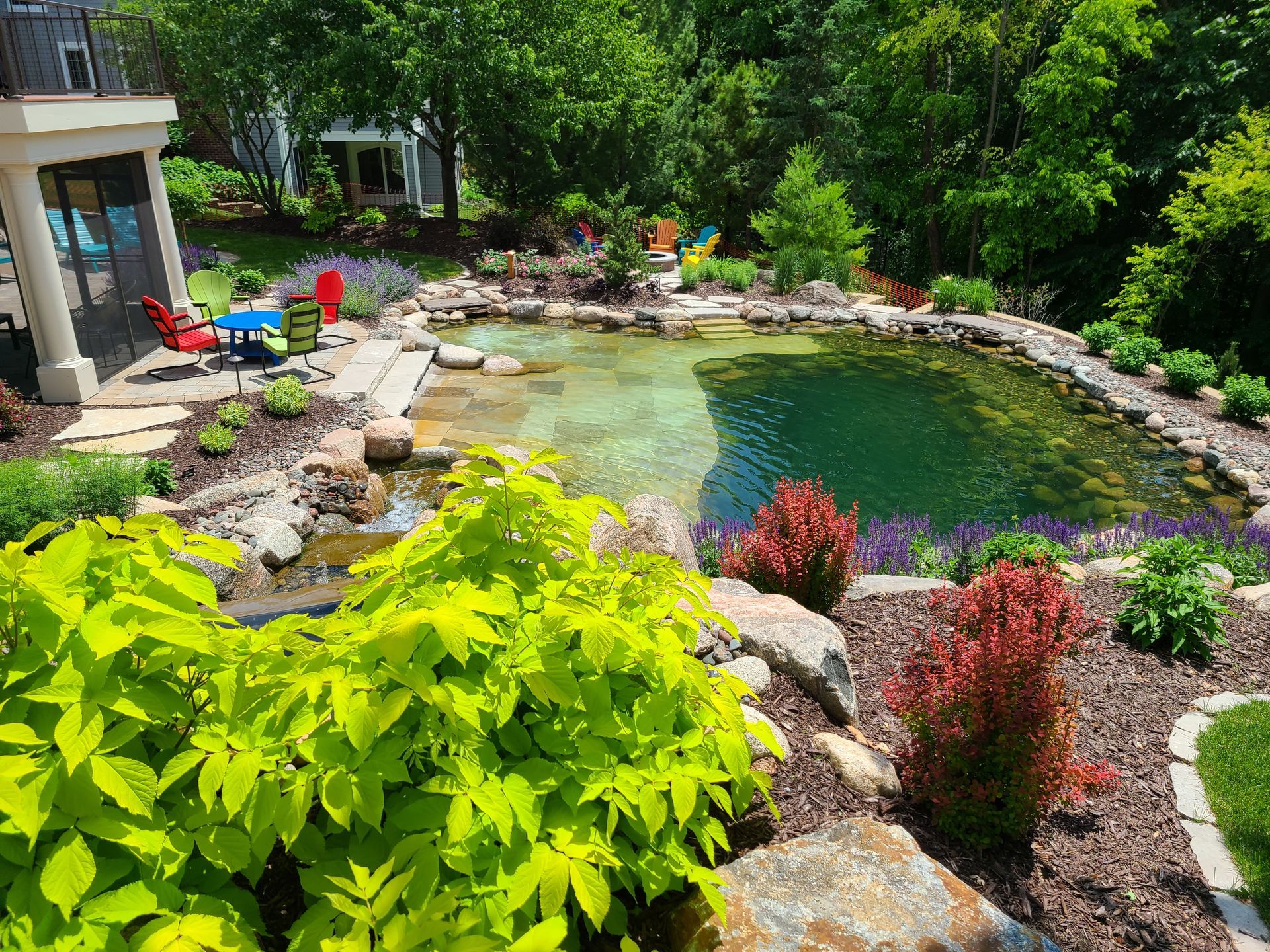 Lush backyard with a natural-looking pond. Green trees surround a multi-level rock border, colorful plants.