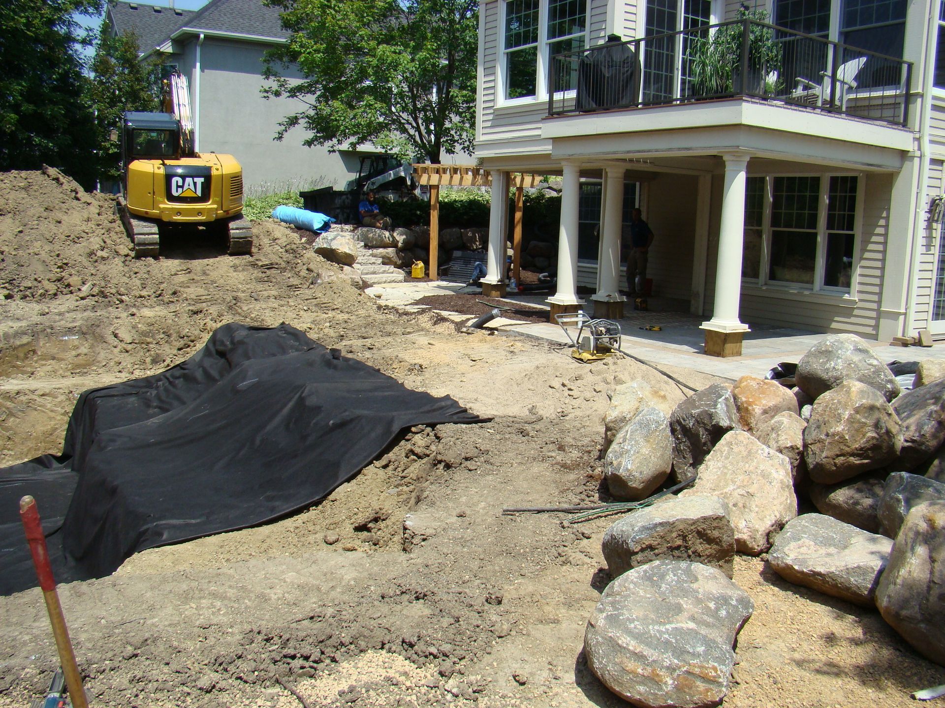 Construction site with excavator, black fabric, large rocks, and a house.