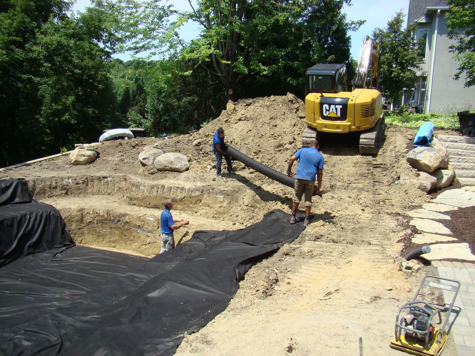 Construction site with workers installing a pond liner, CAT excavator, and large rocks.