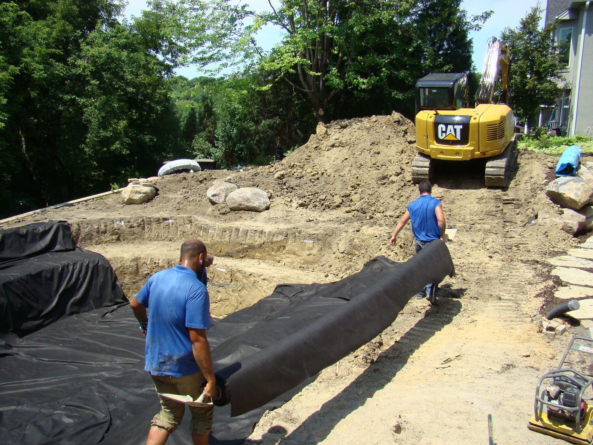 Construction workers installing landscape fabric in a tiered excavation site with a CAT excavator and large rocks.