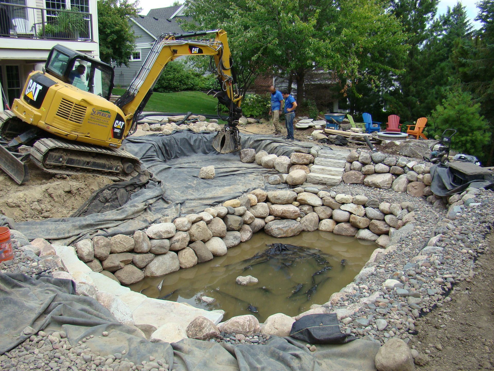 Yellow excavator digging pond, stone walls being built. Two people observe construction in backyard.