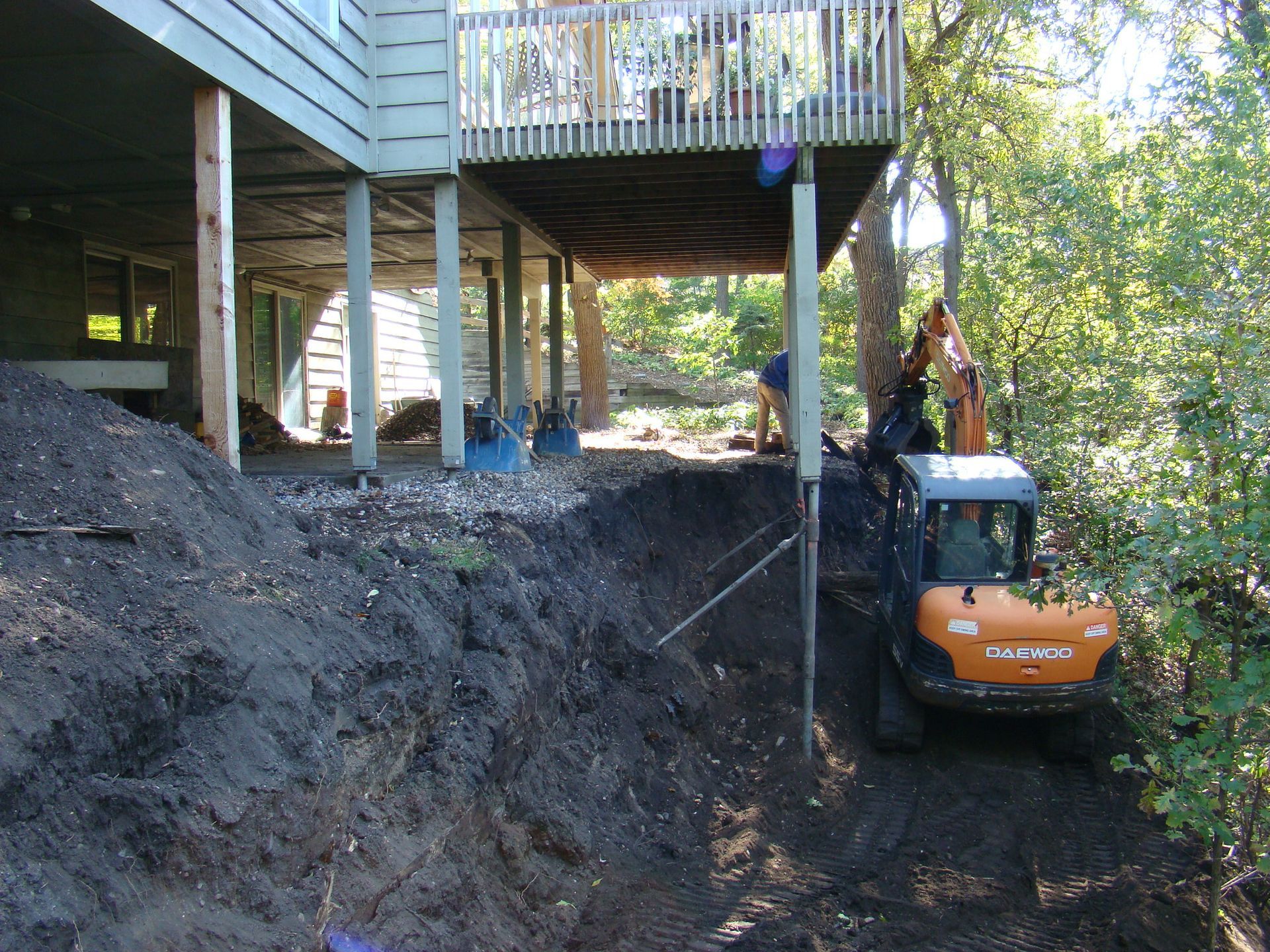 Excavator on a hillside under a raised deck, digging and supporting it.