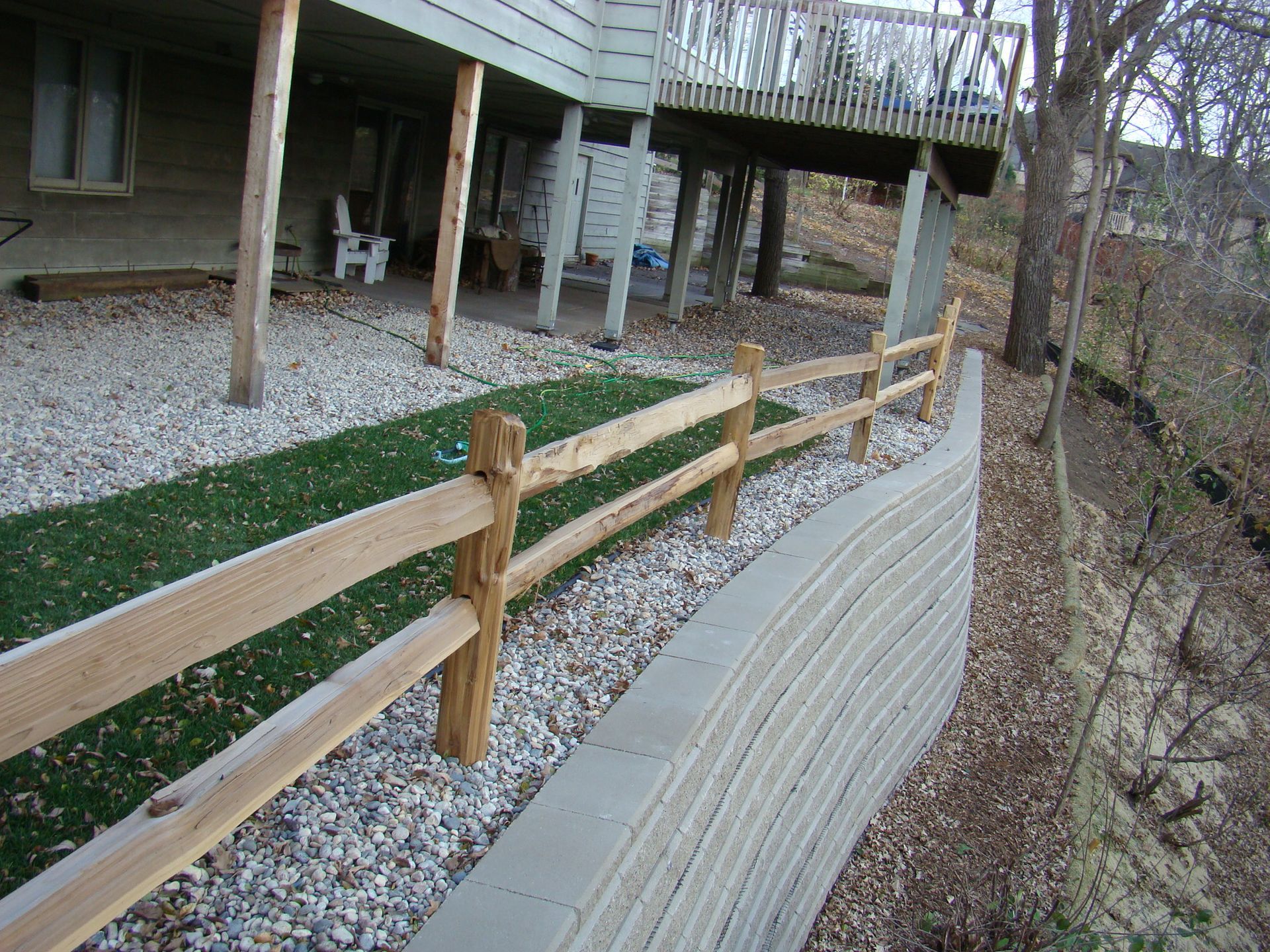Wooden split-rail fence alongside a concrete retaining wall, bordering a grassy area and building's deck.