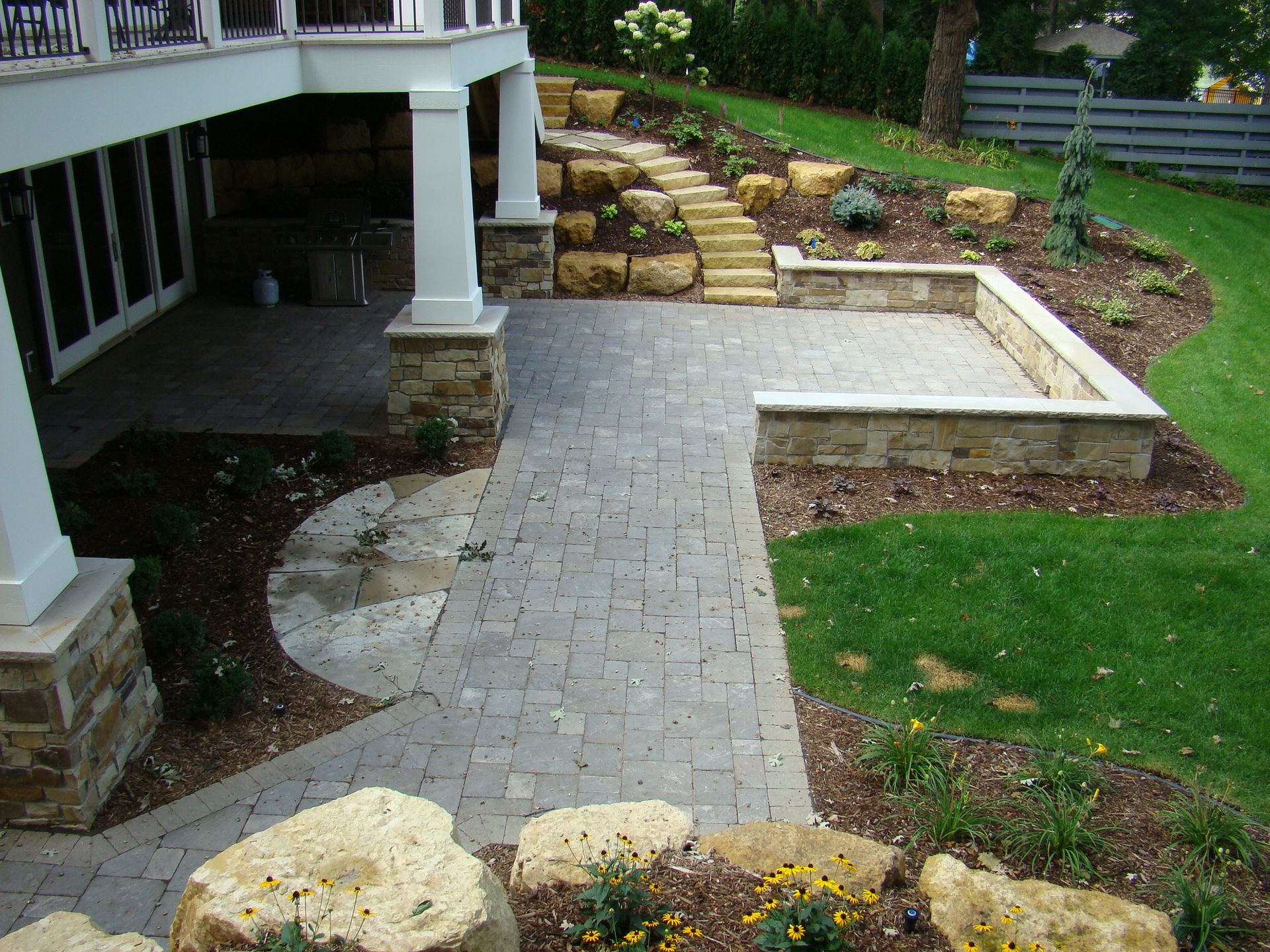 Stone patio and retaining walls with steps, connecting to a grassy yard and tiered landscaping, beneath a home.