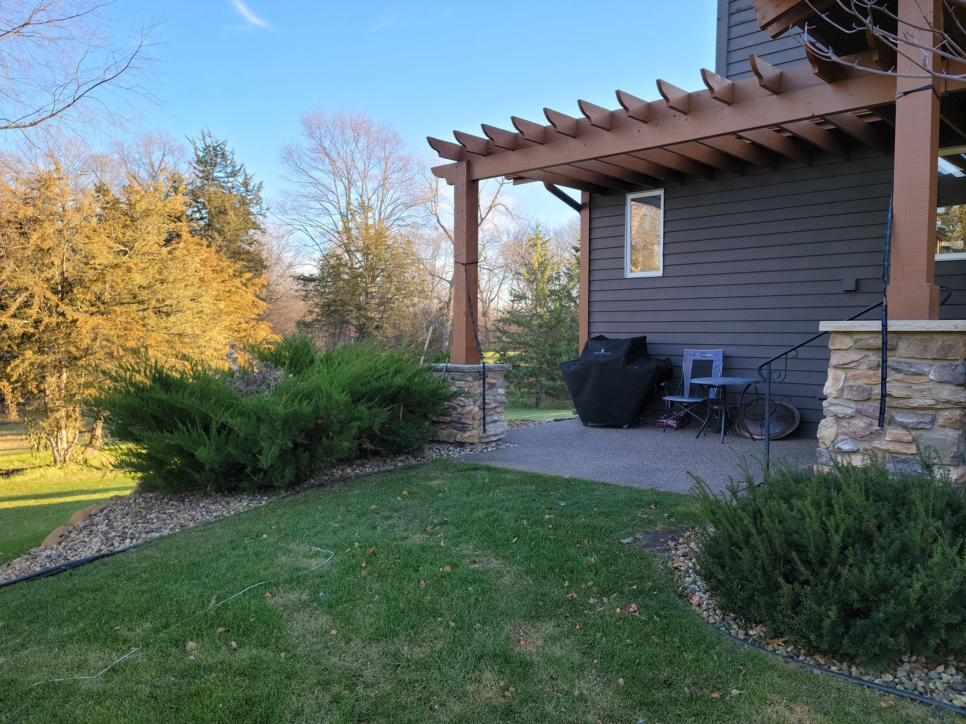 A patio with a dark gray house, pergola, grill, and bushes. Green grass and trees are in the background.