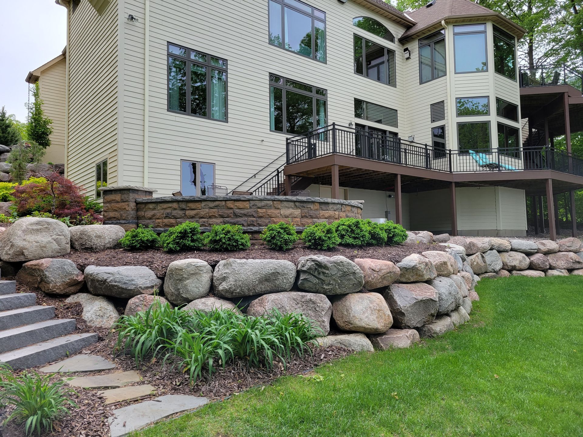 Two-story beige house with stone retaining walls, patio, and deck. Green lawn and shrubs in the foreground.
