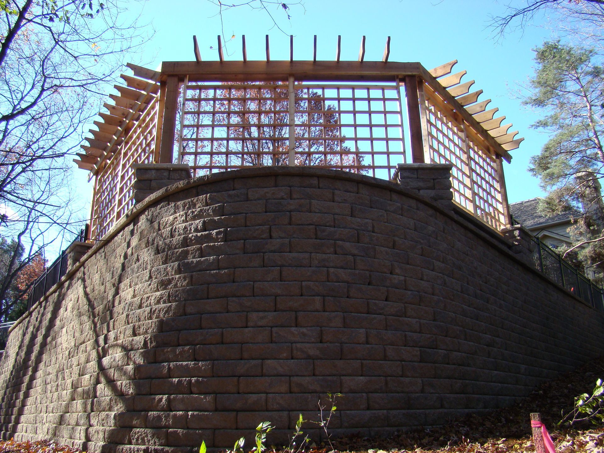 A wooden pergola atop a curved retaining wall made of brown bricks. Sunny, outdoor setting.
