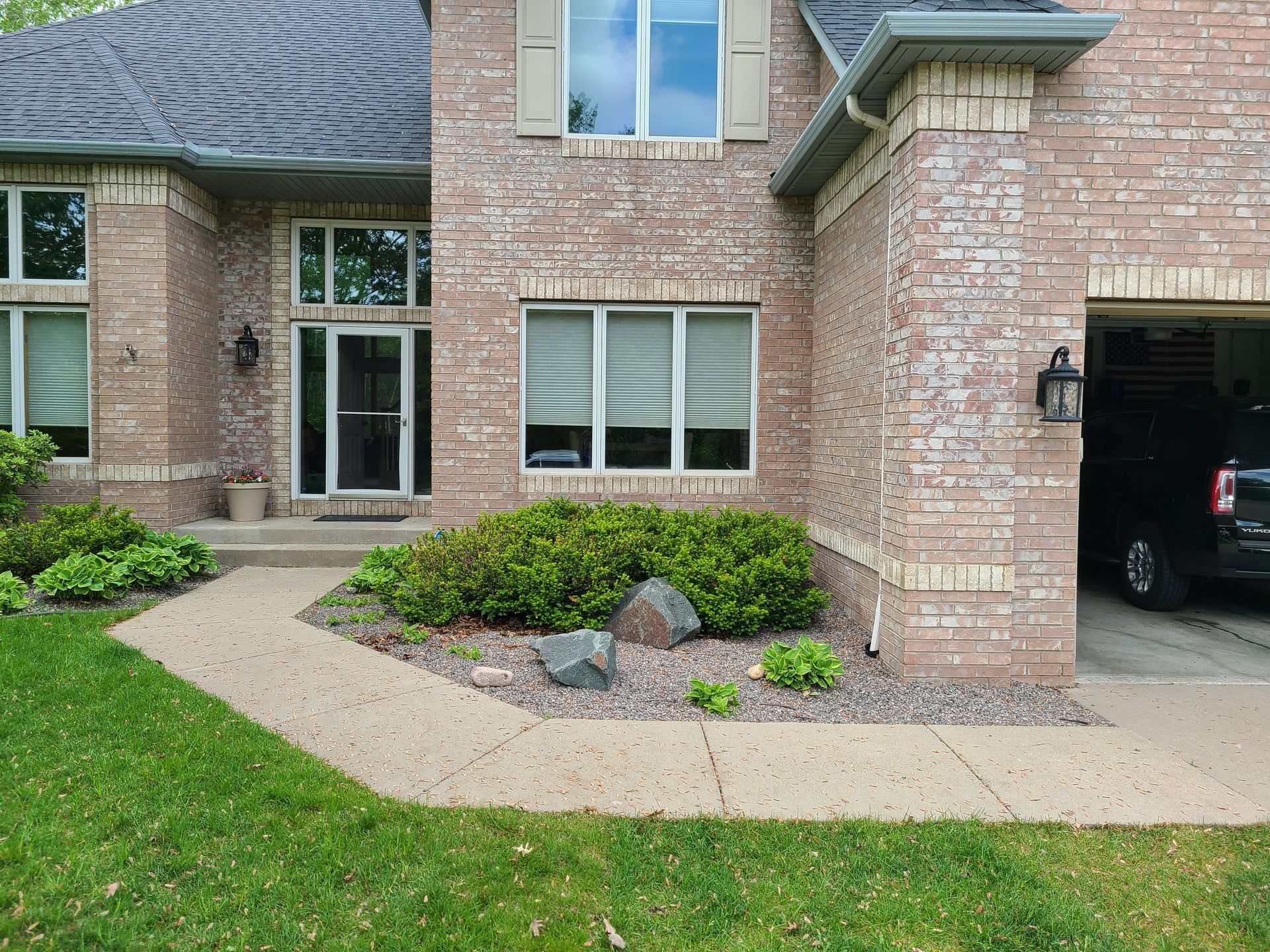 Exterior view of a brick house with a front path, bushes, and a garage with a car parked inside.