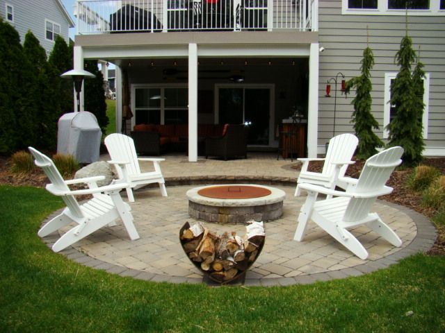 Patio with a fire pit surrounded by white Adirondack chairs, near a house with a deck.