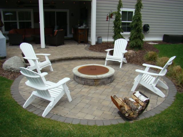 A circular patio with Adirondack chairs around a fire pit, firewood, and backyard greenery.