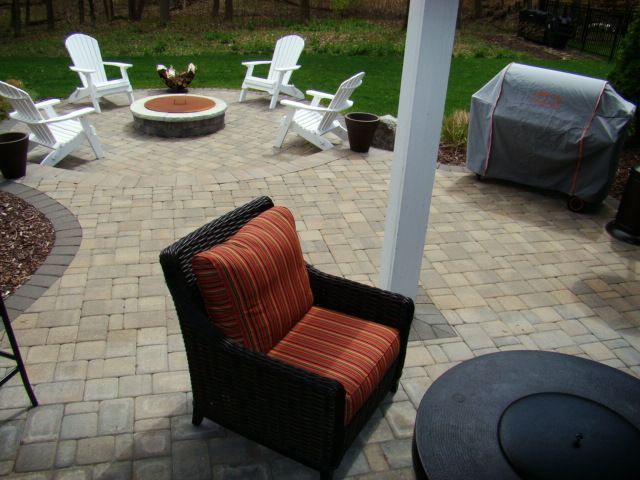 Patio with seating around a fire pit, chair in foreground, grill covered, sunny outdoor setting.