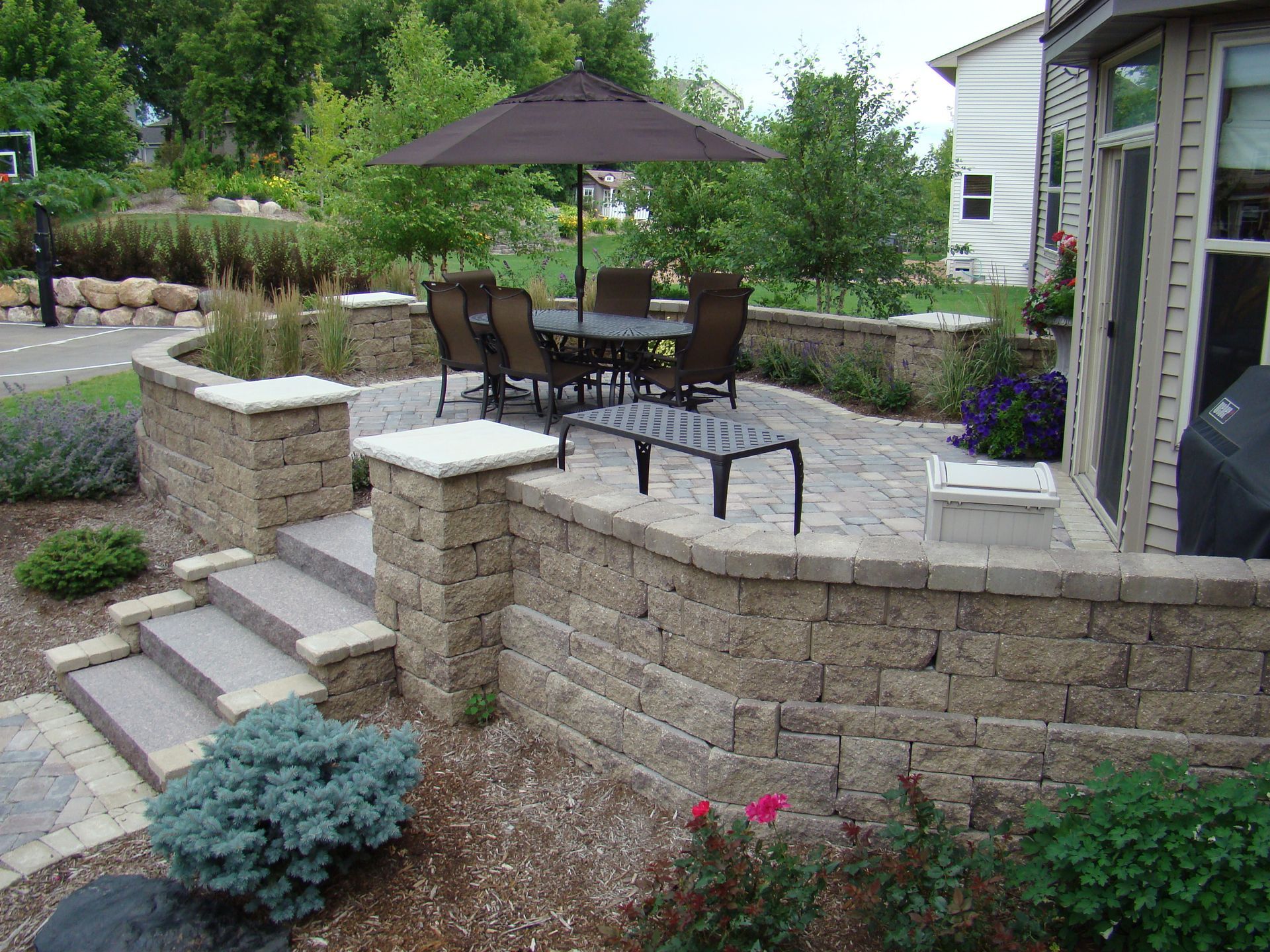 Stone patio with dining set, umbrella, and low retaining walls, steps, and landscaping.
