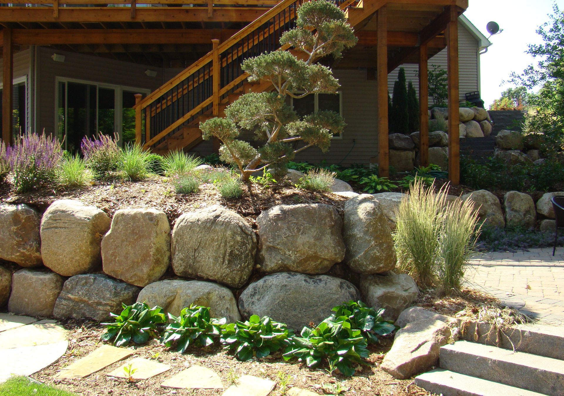 Stone retaining wall with plants and small tree, under a wooden deck.