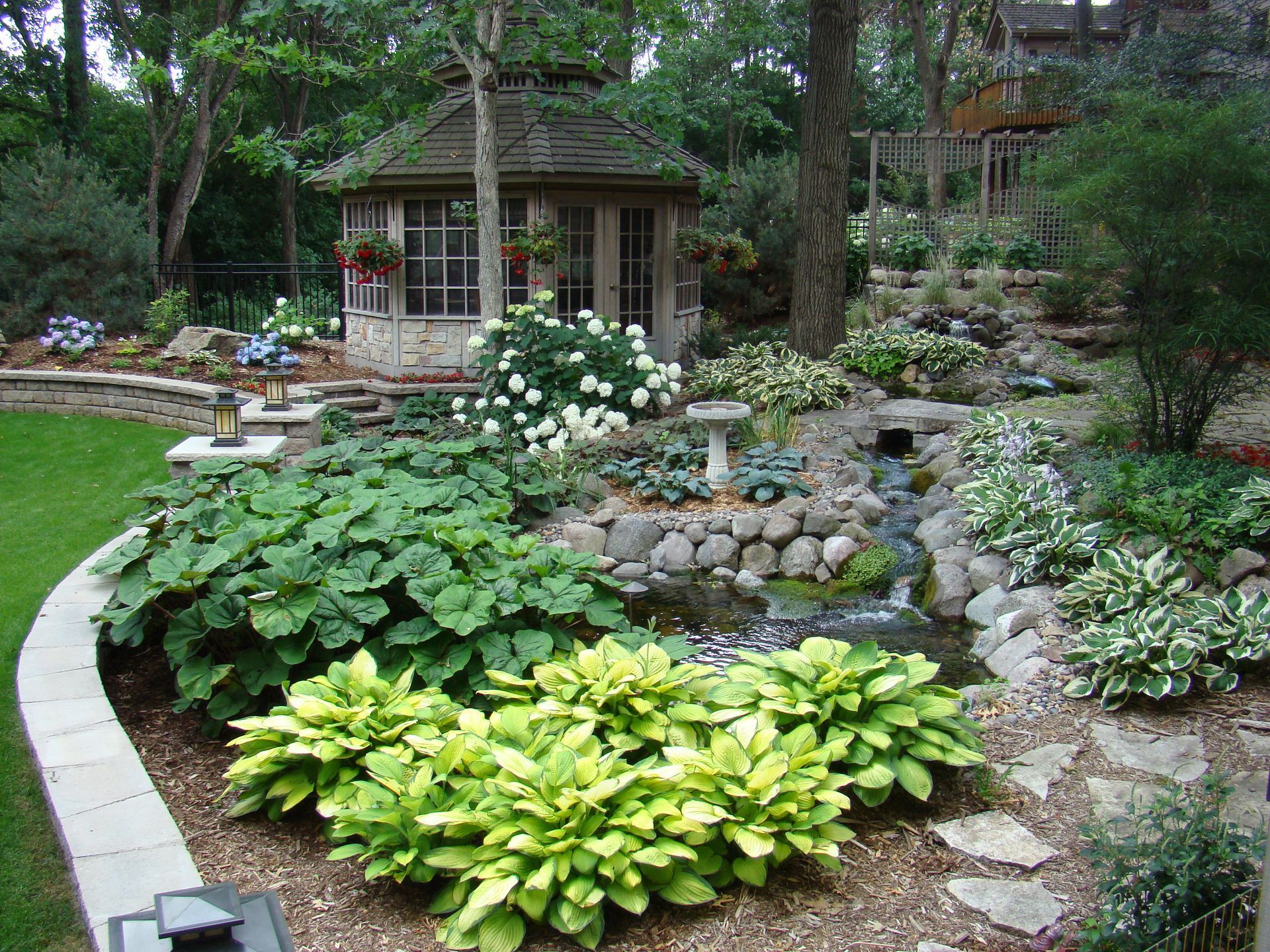 A lush, tiered garden with a small gazebo, pond, and various green and white plants.