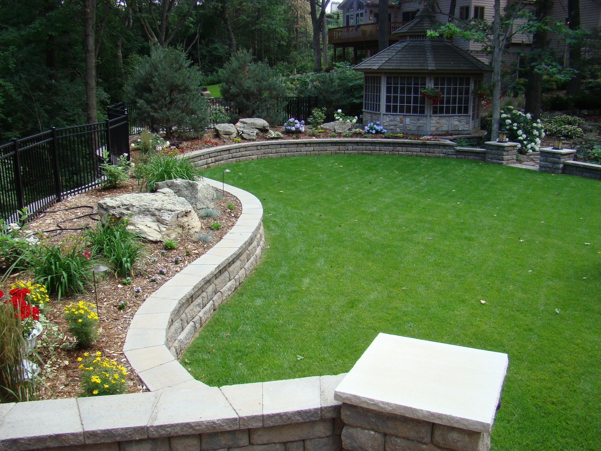 Landscaped backyard with lawn, curved stone retaining wall, and gazebo.
