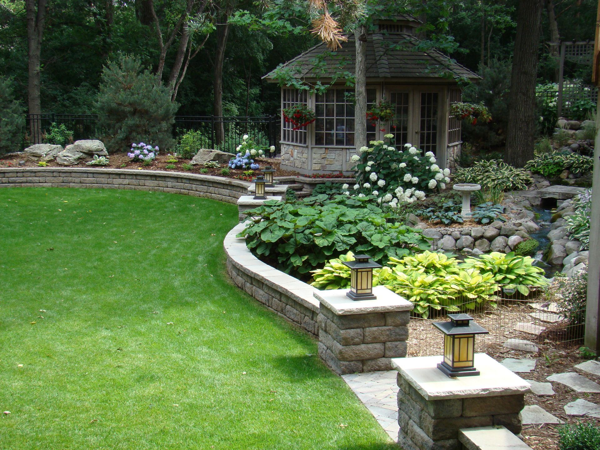 Lush backyard with tiered landscaping, gazebo, retaining wall, and green lawn. Lanterns on pillars.