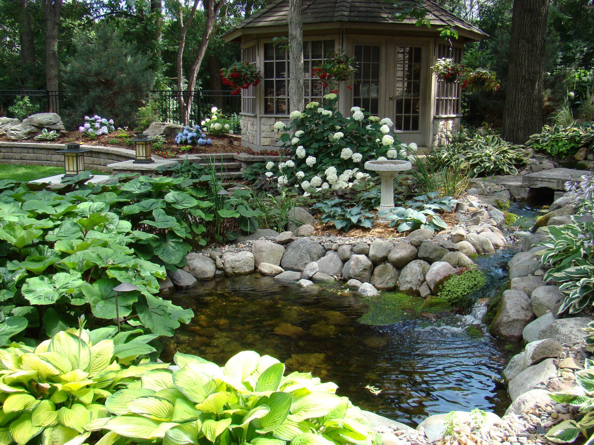A garden scene with a pond, a gazebo, and various plants and flowers.