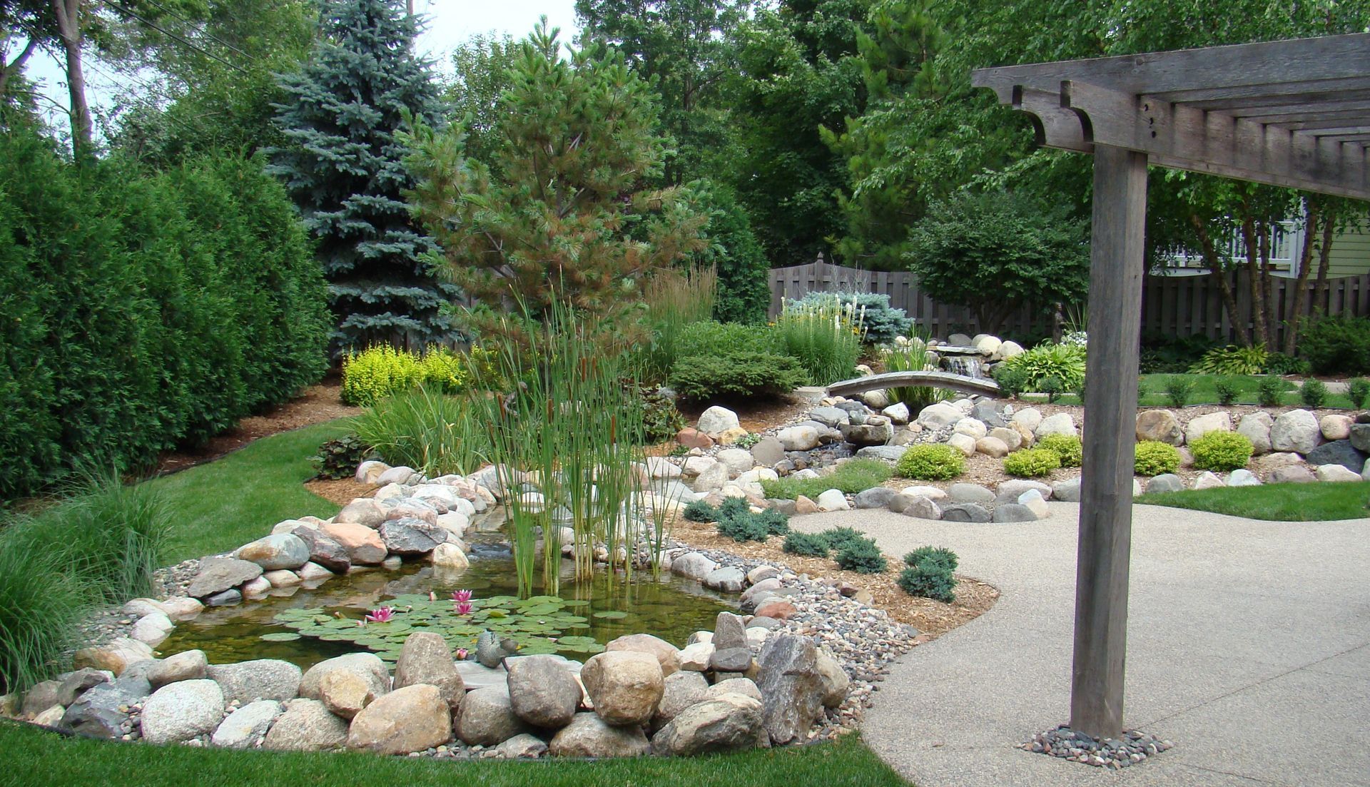 A landscaped backyard with a pond, rocks, plants, and a pergola.