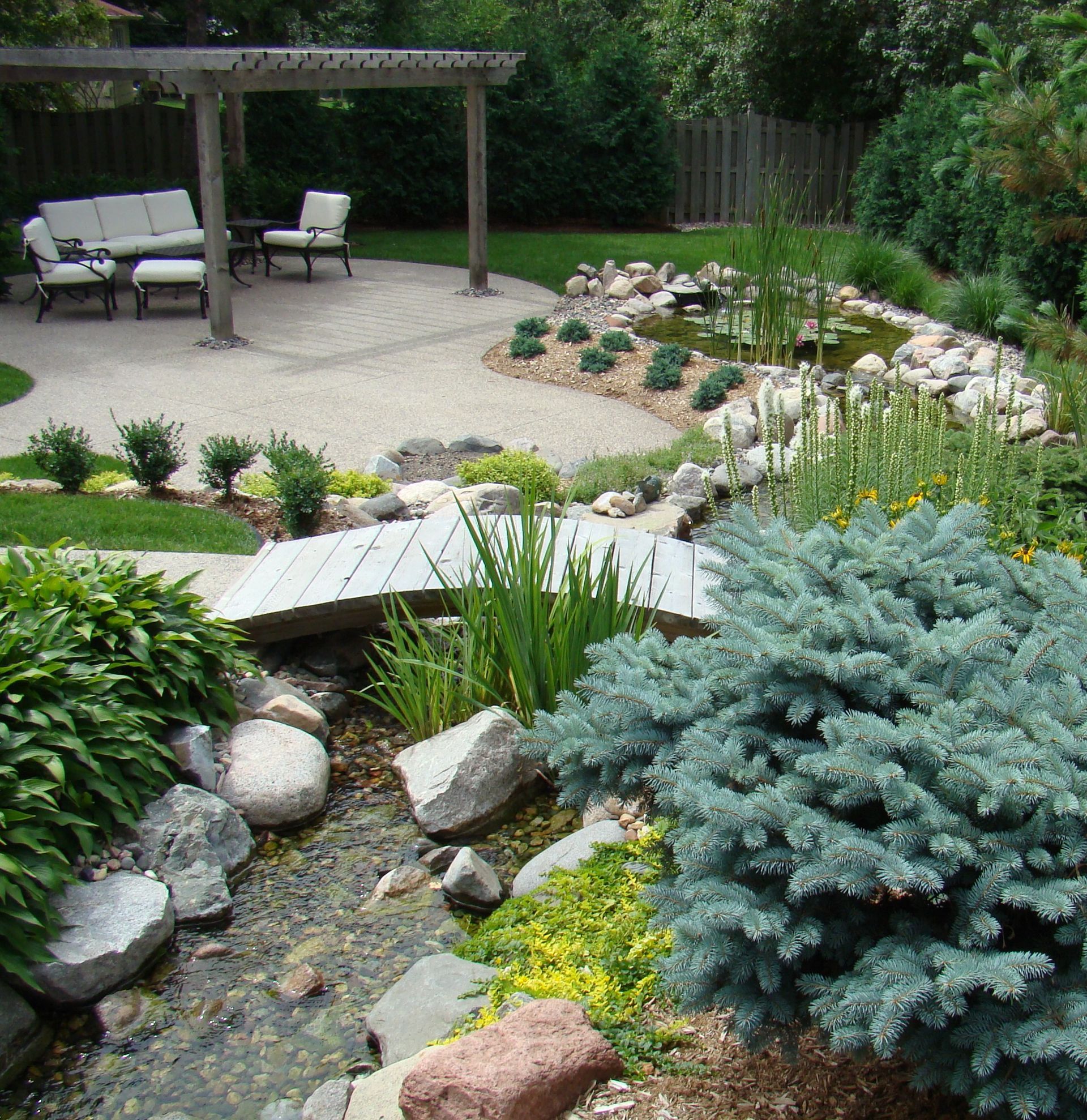 Backyard with stone patio, water features, bridge, and seating area under a pergola.