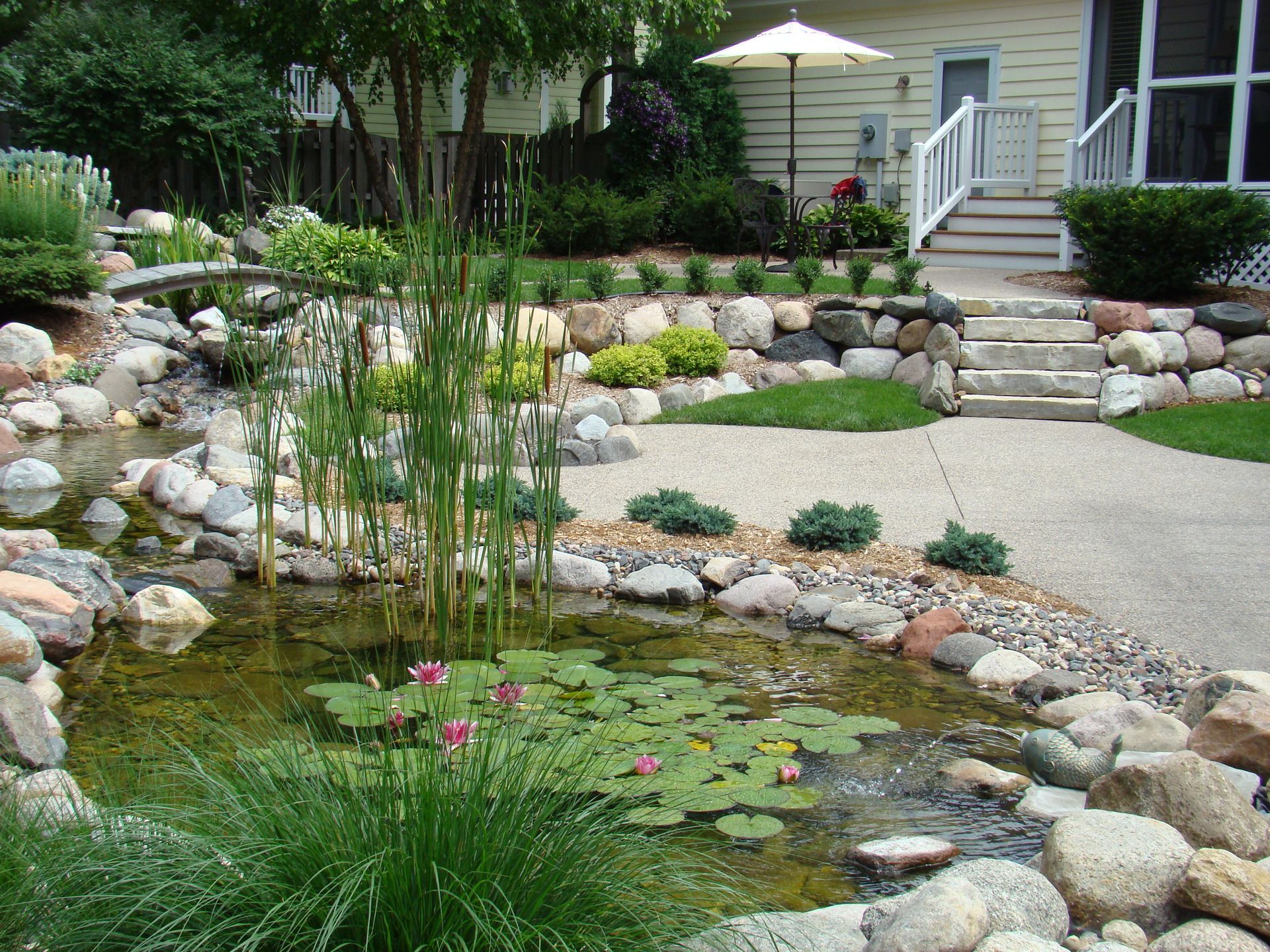 Landscaped backyard with pond, rocks, and small bridge leading to a home with patio steps.