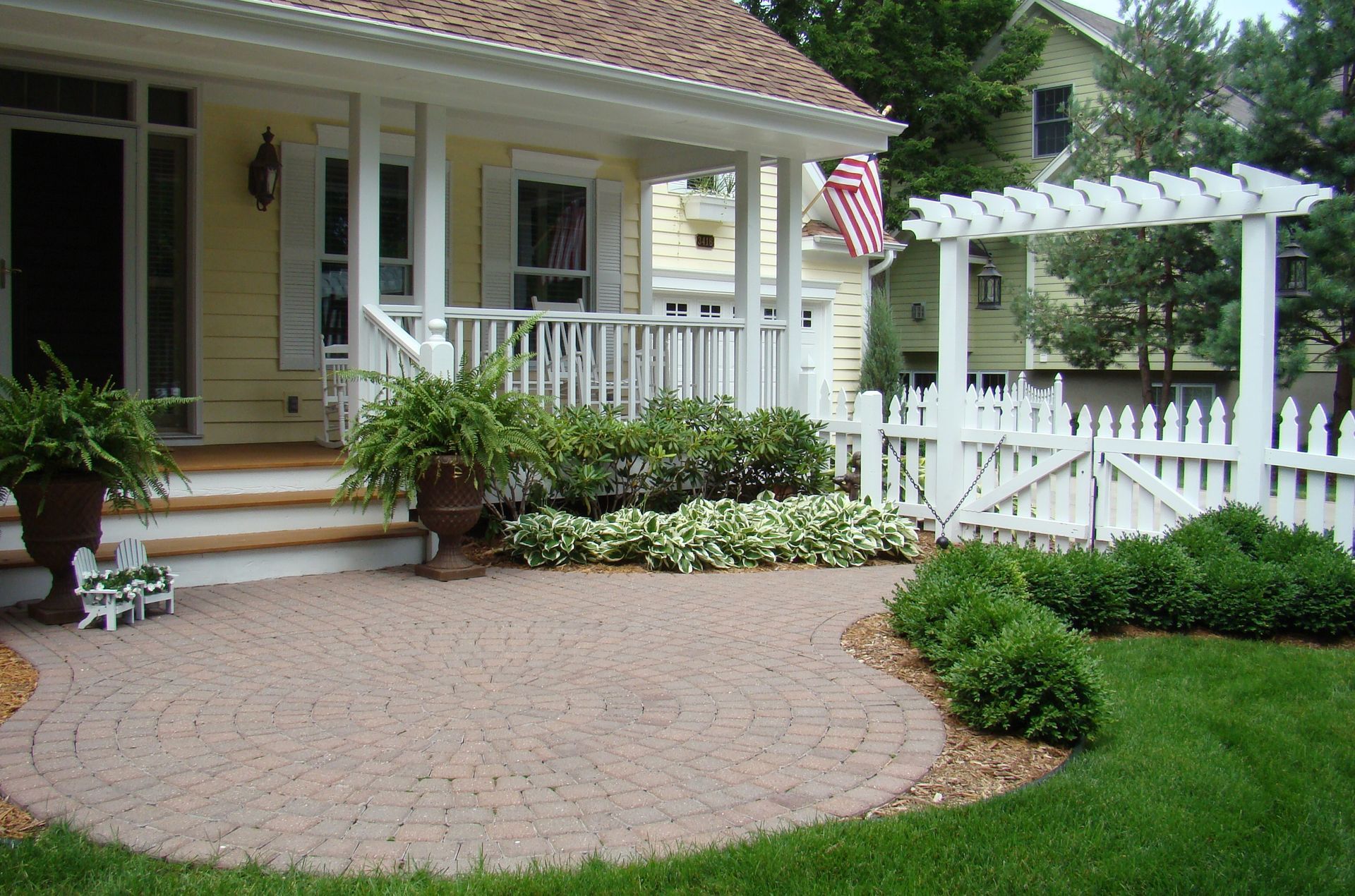 Yellow house with porch, brick patio, white picket fence, and green lawn.