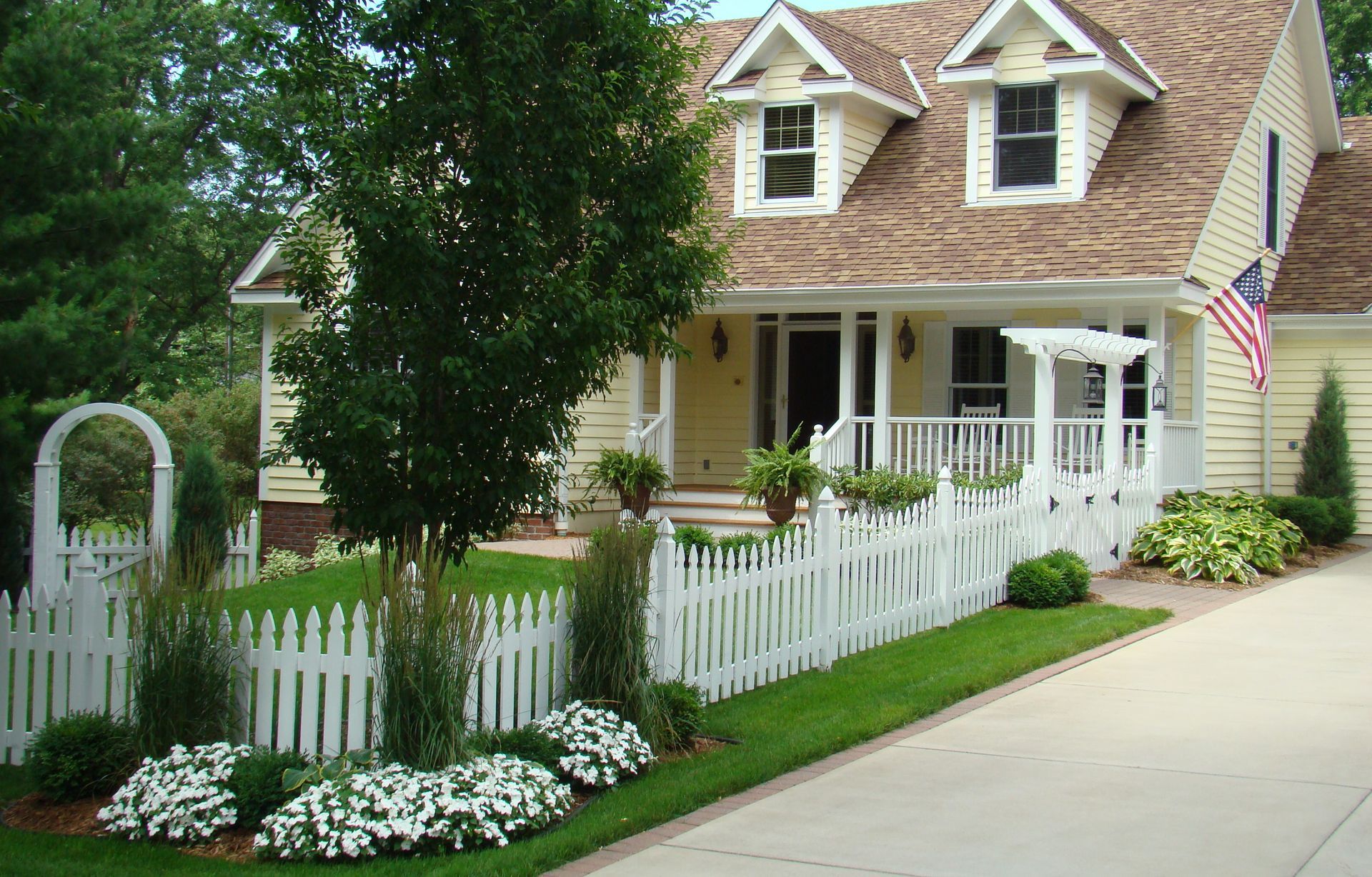 Yellow house with white picket fence, front porch, and American flag; green lawn, tree.