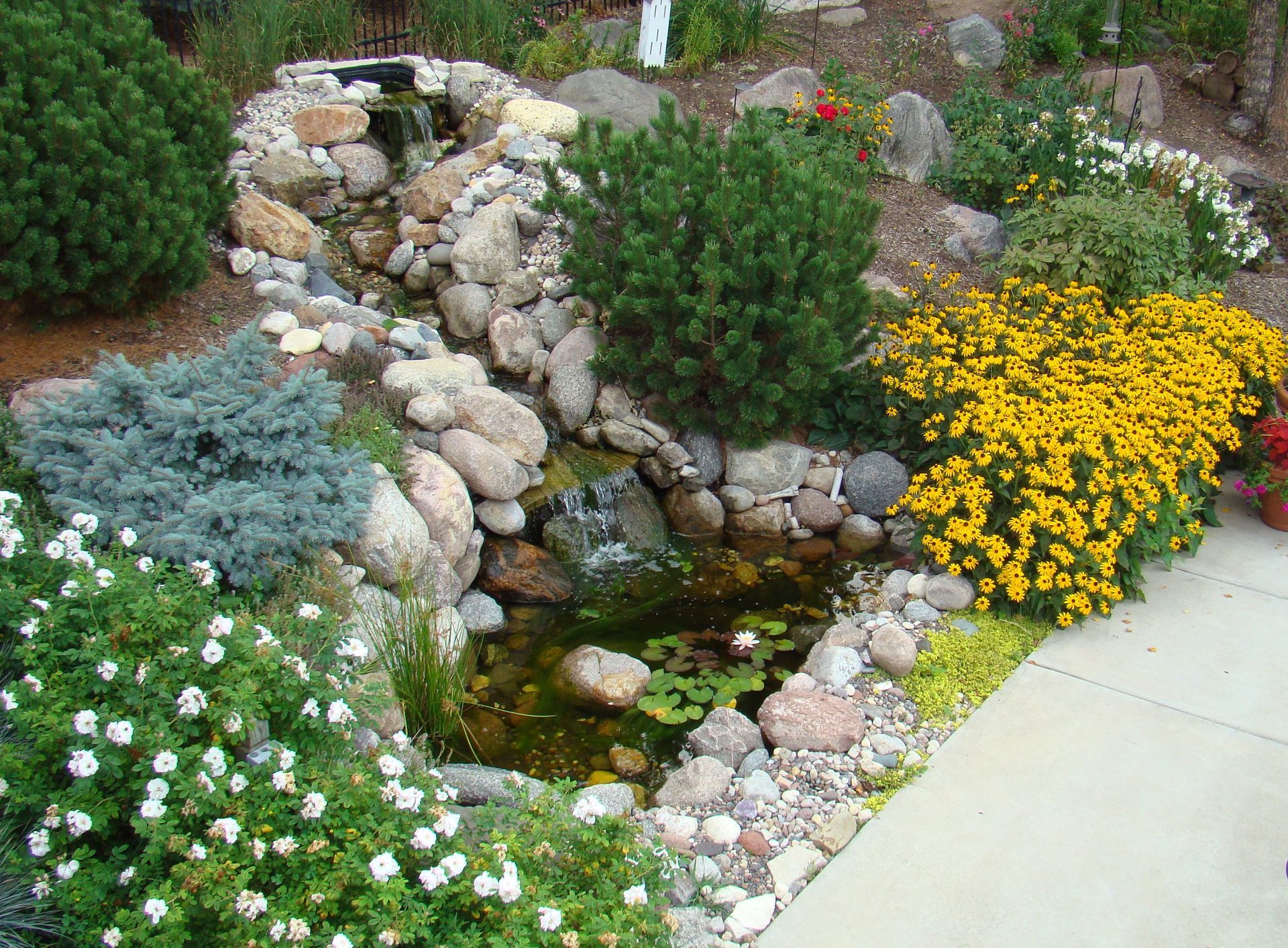 A small pond with a rocky waterfall surrounded by greenery and colorful flowers.