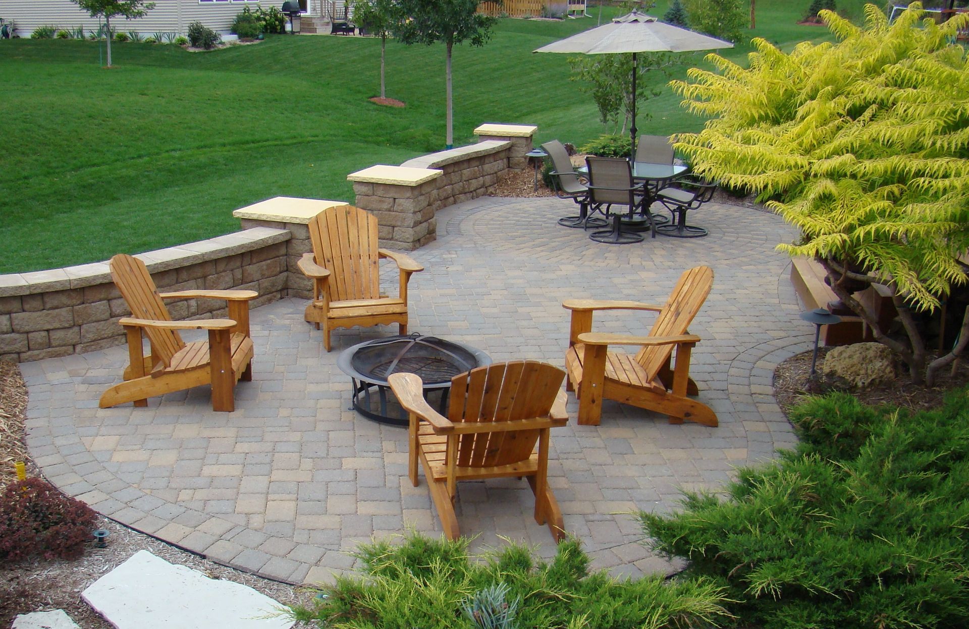 Stone patio with Adirondack chairs around a fire pit and a table with chairs under an umbrella, surrounded by greenery.