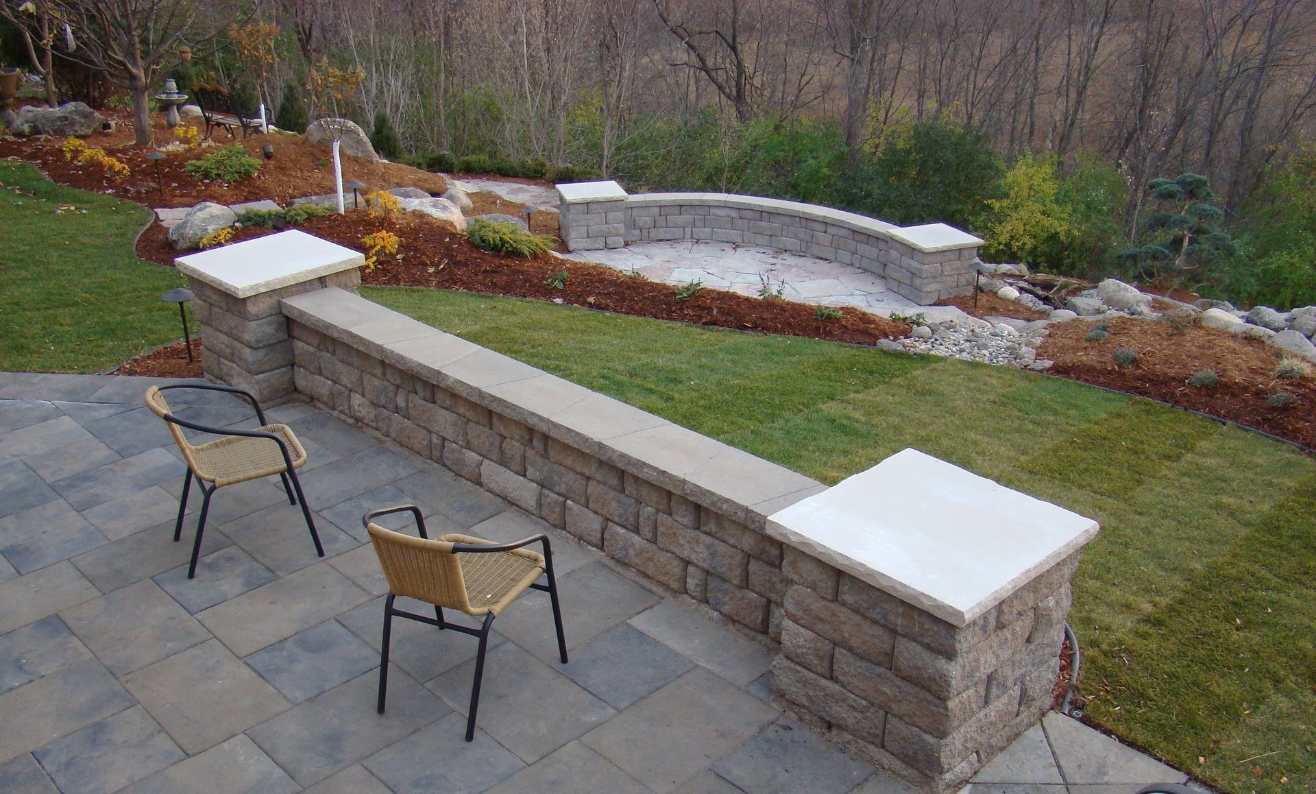 Patio with stone wall, chairs, and garden amphitheater; green lawn, brown mulch, and trees in background.
