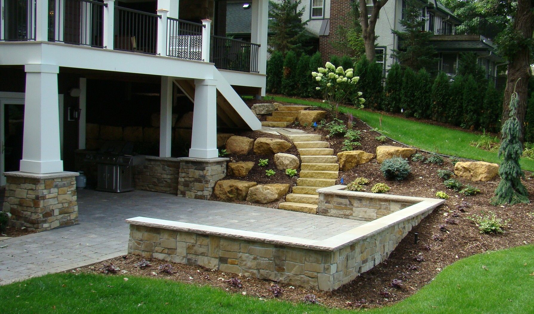 Stone patio under a multi-level deck, with stone steps leading to a higher level with landscaping.