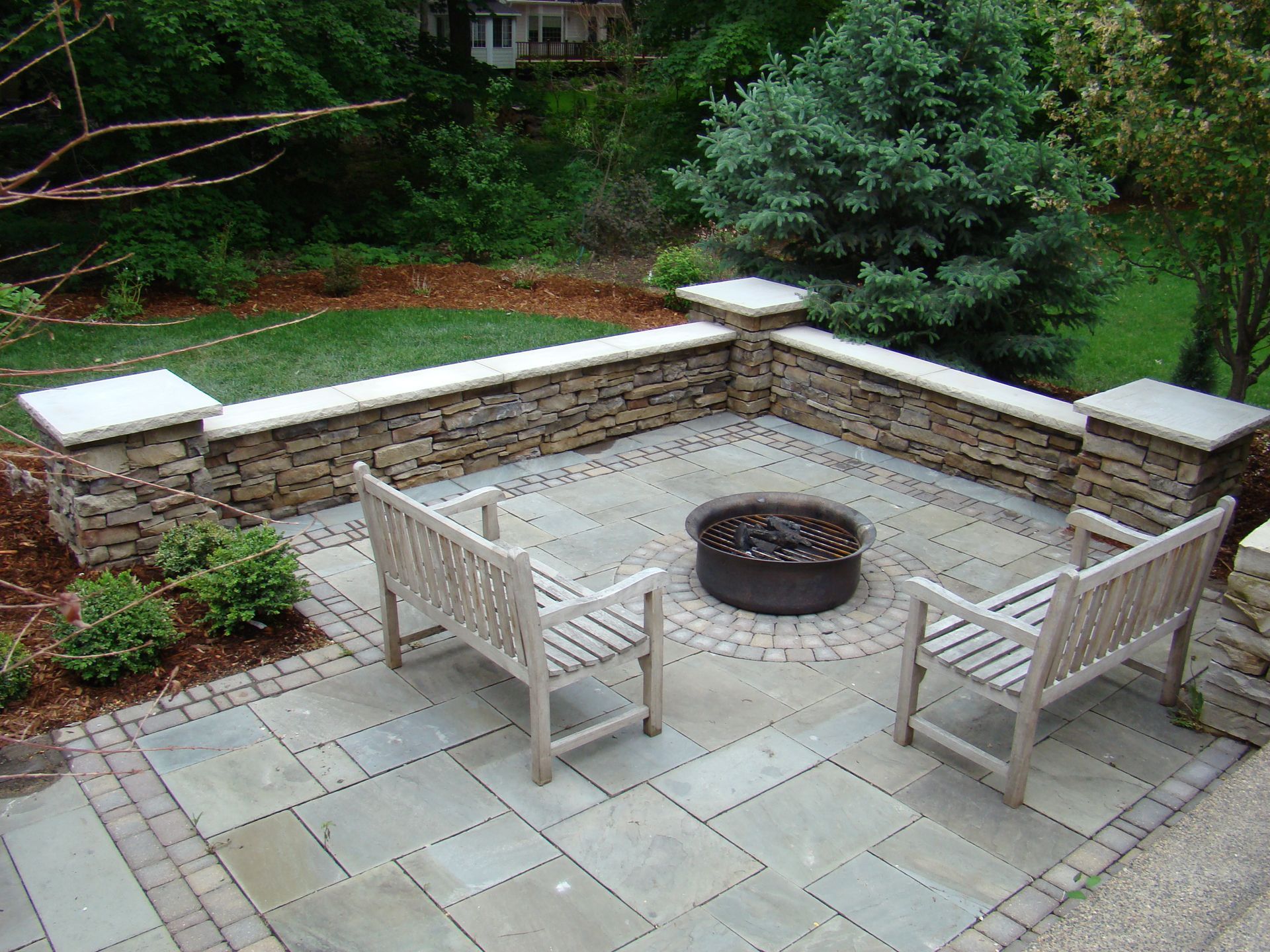 Patio with stone wall, benches, and fire pit surrounded by green grass and trees.