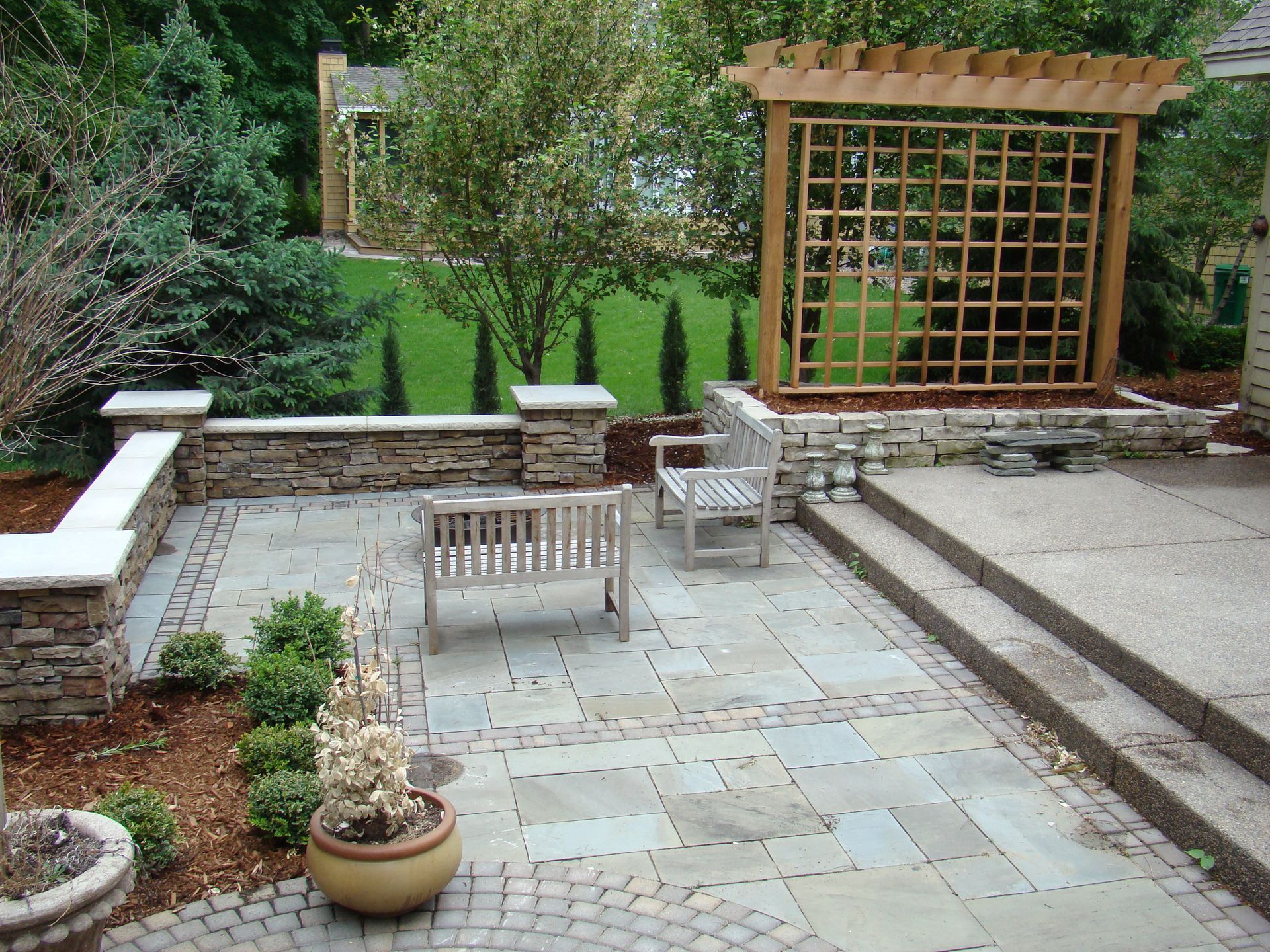 Stone patio with wooden benches, trellis, and low stone walls surrounded by greenery.