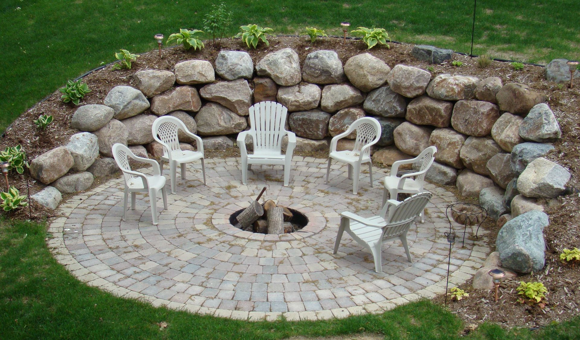 Stone-walled fire pit with a circular brick patio, surrounded by plastic chairs on grass.