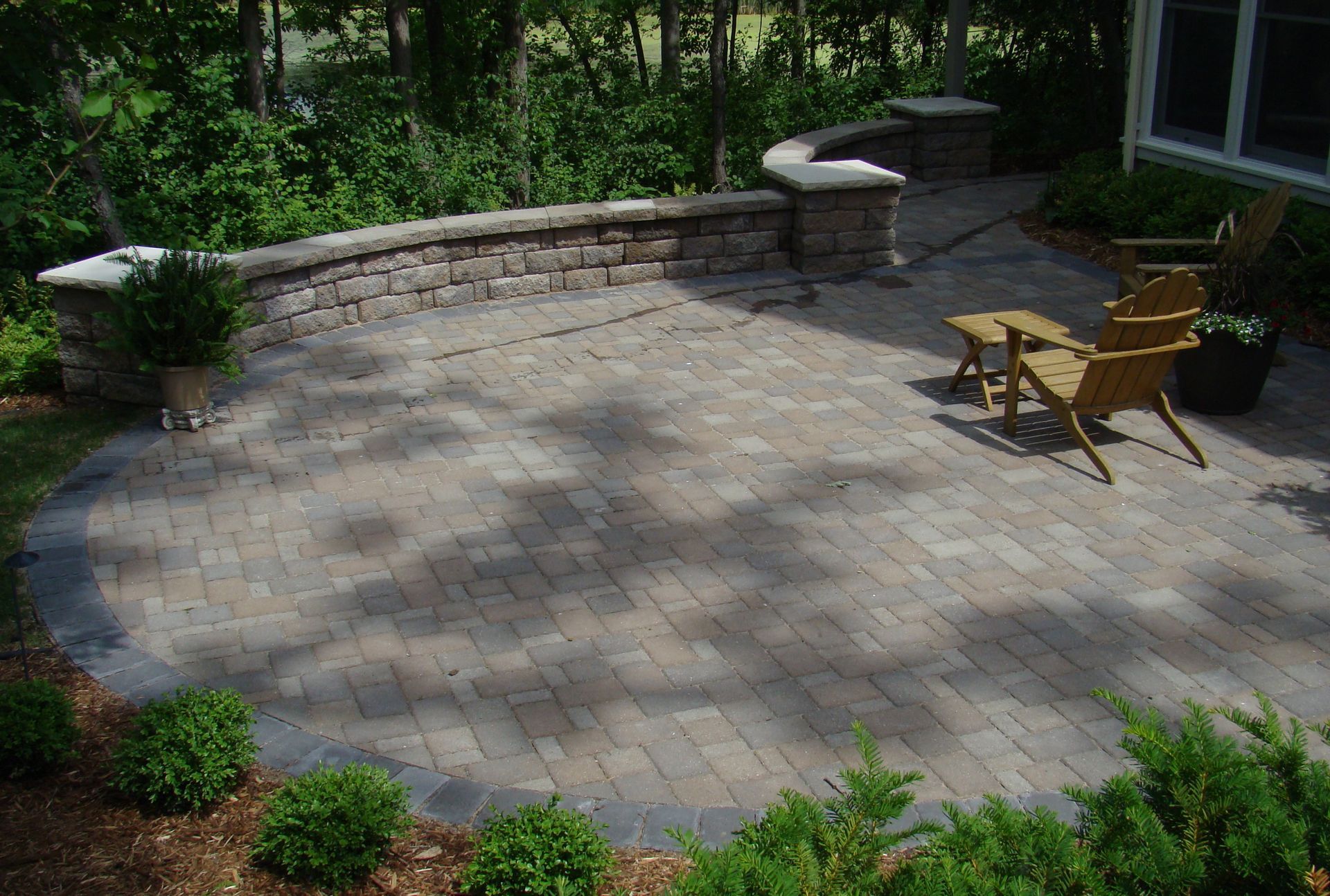 Brick patio with seating and retaining wall, surrounded by shrubs and trees.