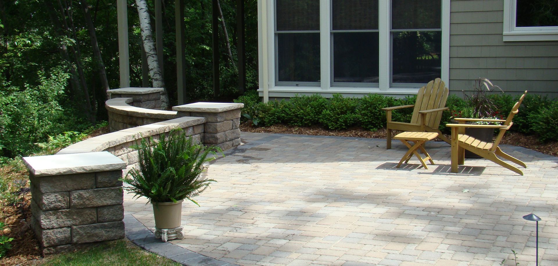 Patio with stone wall, two wooden chairs, and potted plant, next to a house and greenery.
