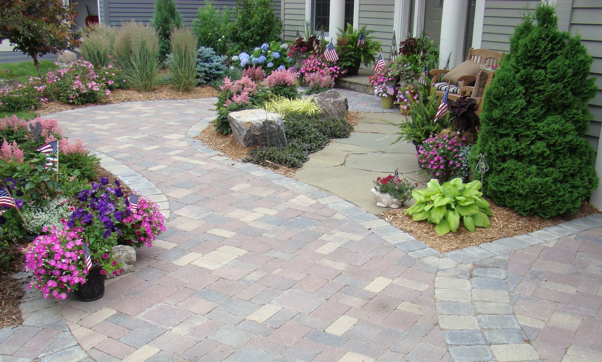 Brick pathway curves through a colorful garden, leading to a light-colored house.
